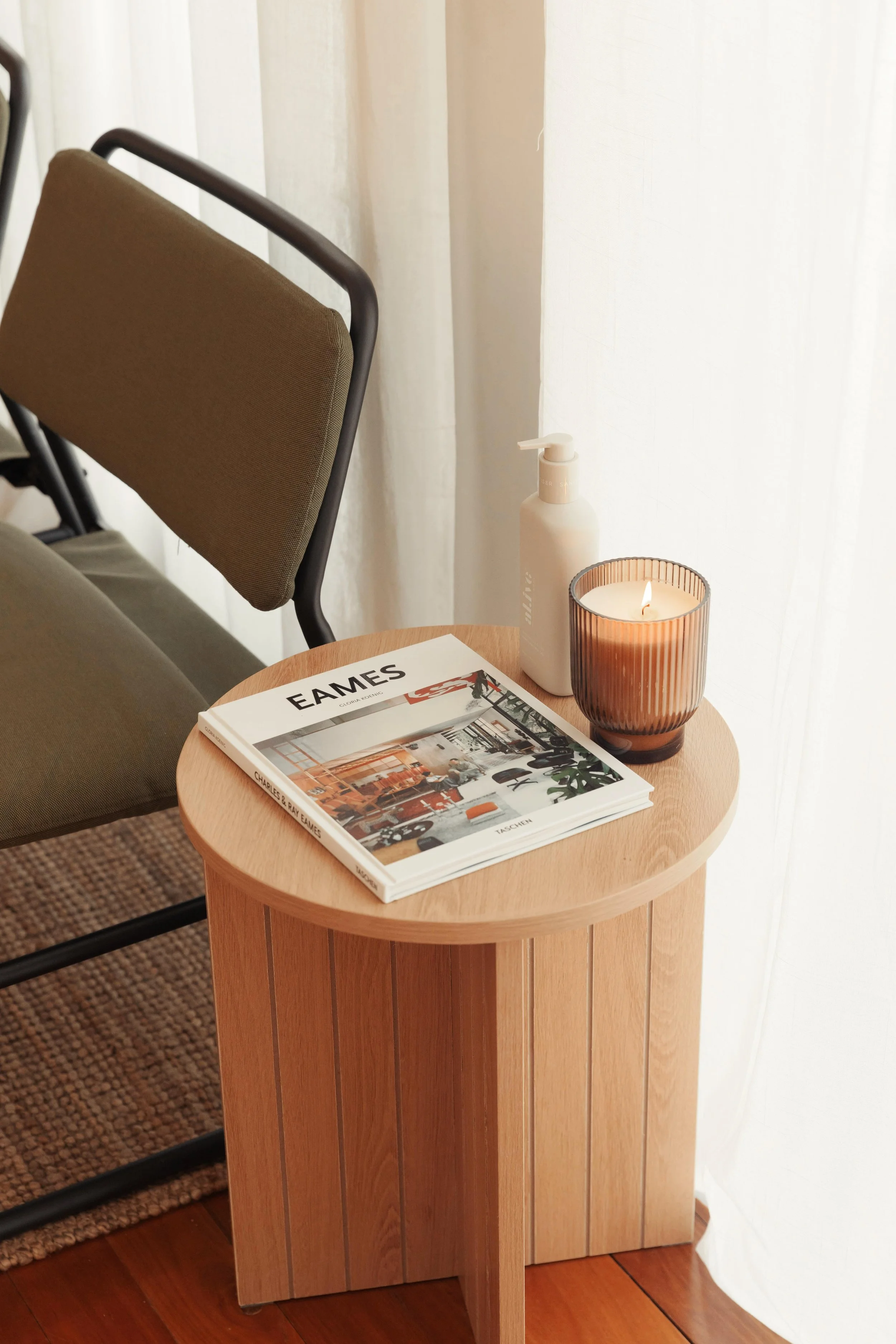 A wooden side table with a magazine, a lamp, and a bottle of lotion next to a chair with a brown cushion and a metal frame, in front of white curtains