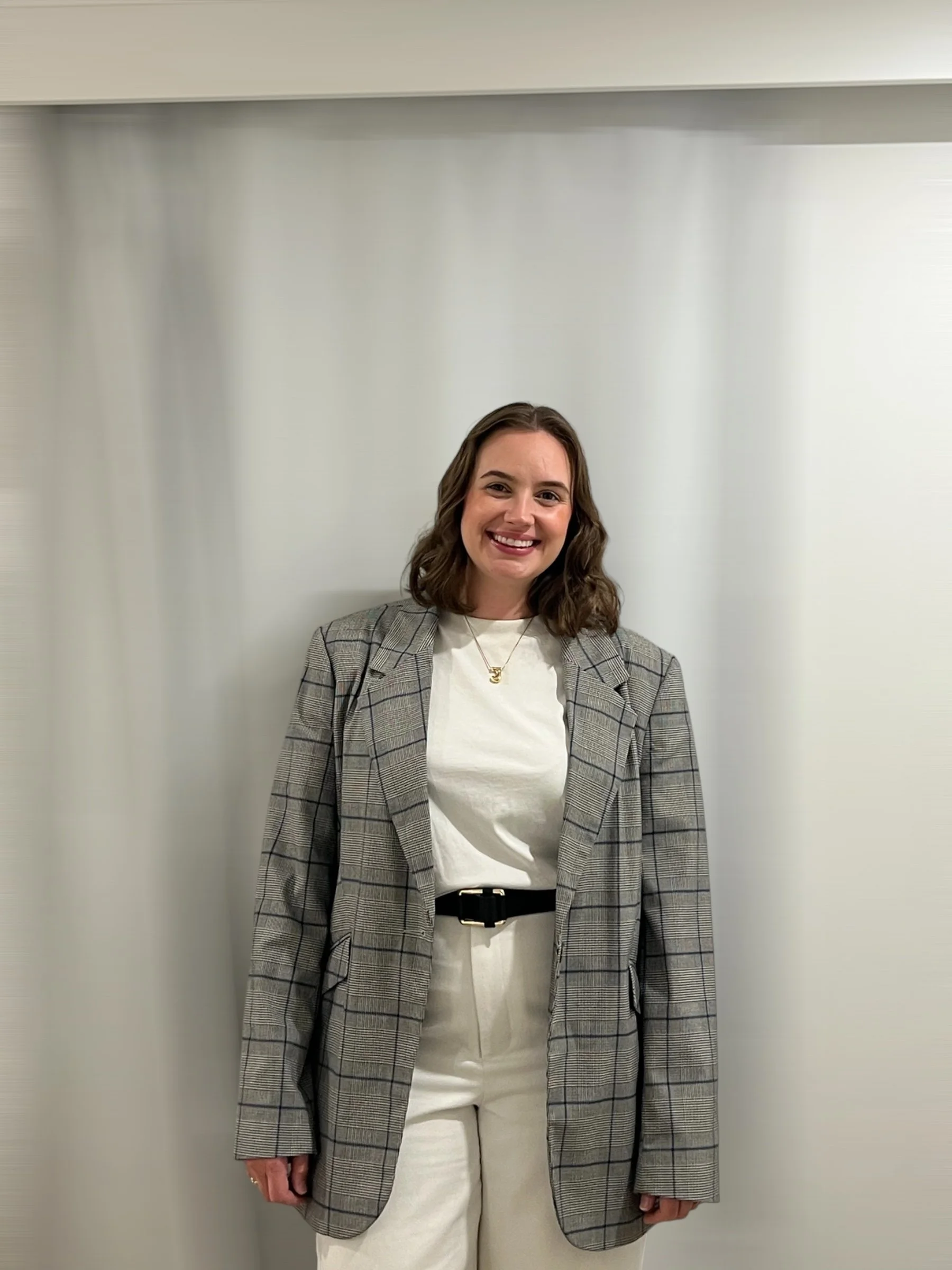 A woman with shoulder-length brown hair and a wide smile, wearing a gray plaid blazer over a white top, standing against a plain light-colored wall.
