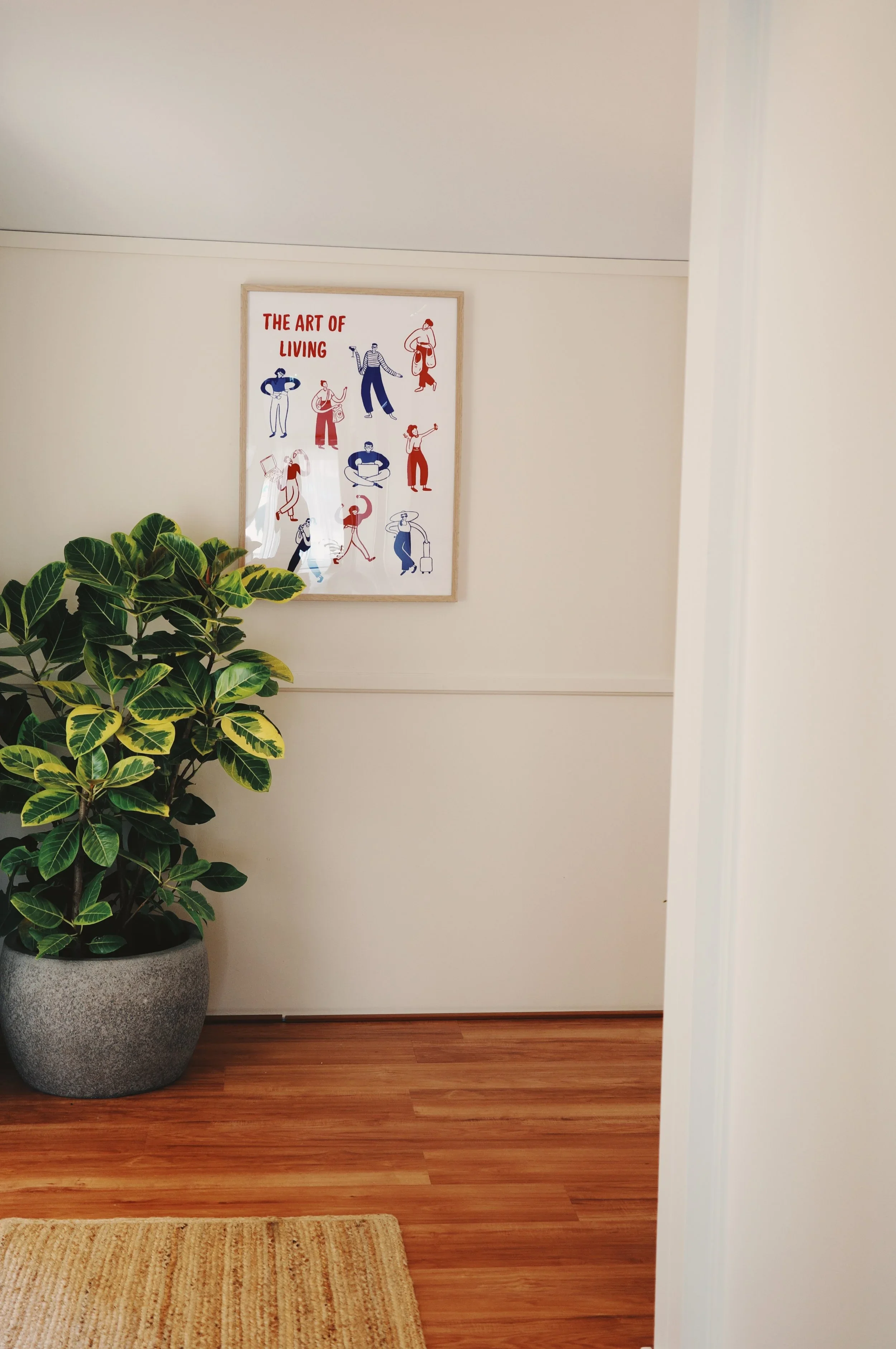 Interior view of a room with a potted plant, a framed art piece on the wall titled 'The Art of Living' with illustrations of people, wooden flooring, and part of a beige rug.