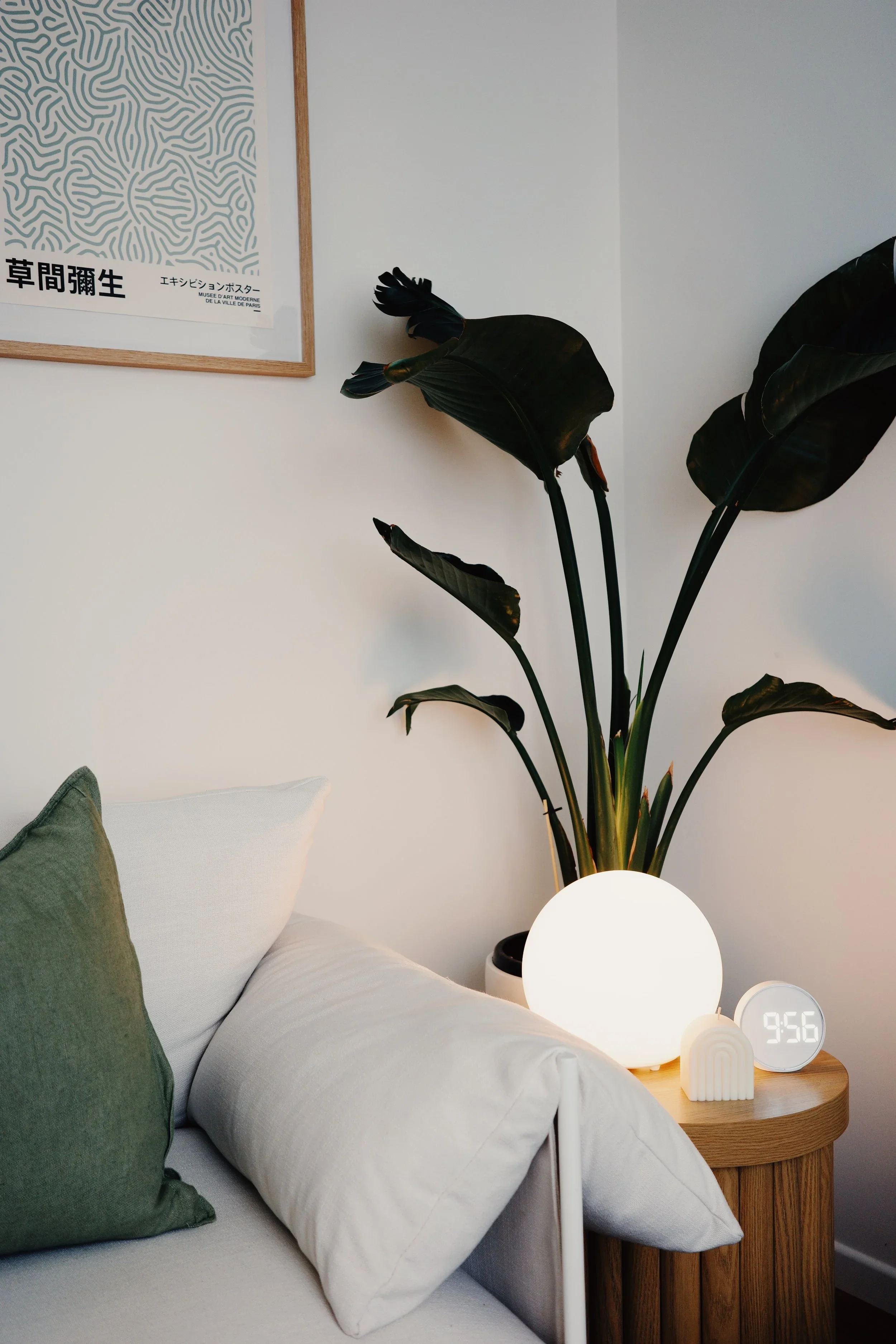 A bedroom with a wooden nightstand featuring a glowing white spherical lamp, an alarm clock displaying 9:56, and two small decorative items. A large dark green plant with broad leaves is next to the nightstand. Part of a white and green bed with pillows is visible, and a framed abstract art print hangs on the wall above.