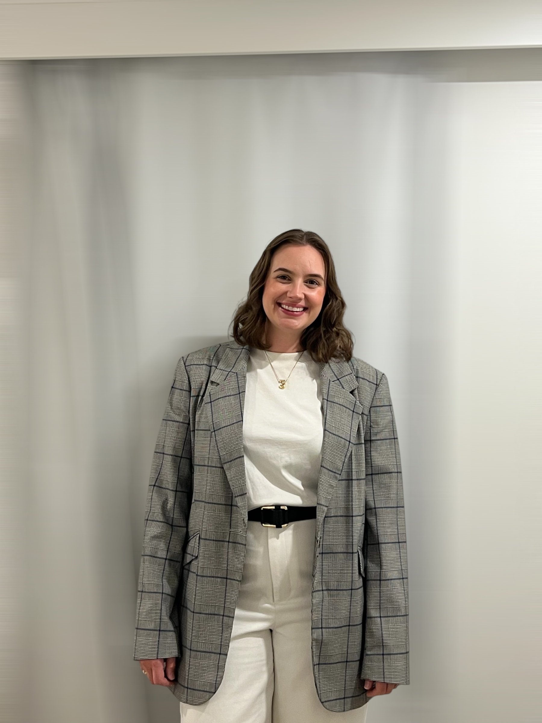 A woman with shoulder-length brown hair, wearing a gray plaid blazer over a white shirt with a necklace, smiling, standing against a plain light-colored wall.