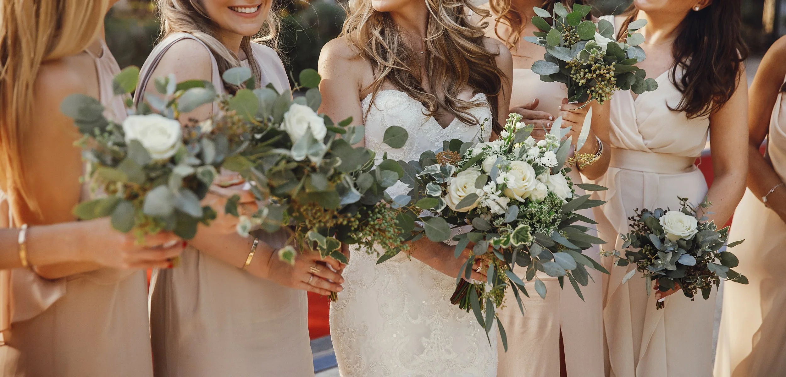 Grupo de mujeres en vestido de novia y vestido de dama de honor, sosteniendo ramos de flores en una ceremonia de boda al aire libre.