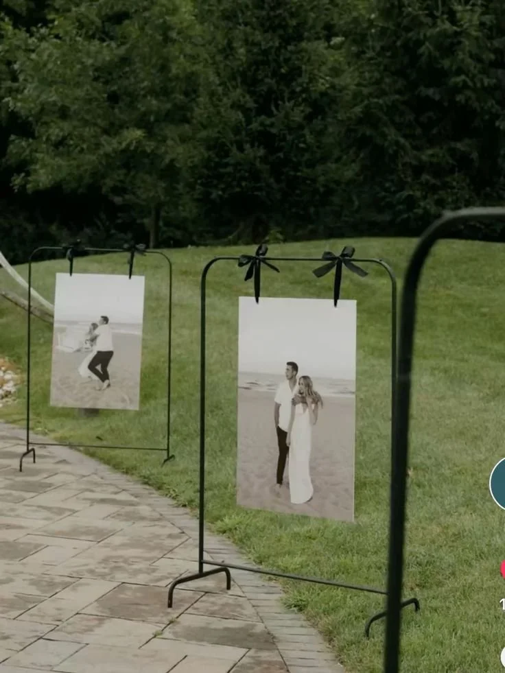 Exposición de fotografías en exteriores, con imágenes en blanco y negro de una pareja en diferentes poses, colgadas en soportes con lazos decorativos en un parque con césped y árboles.