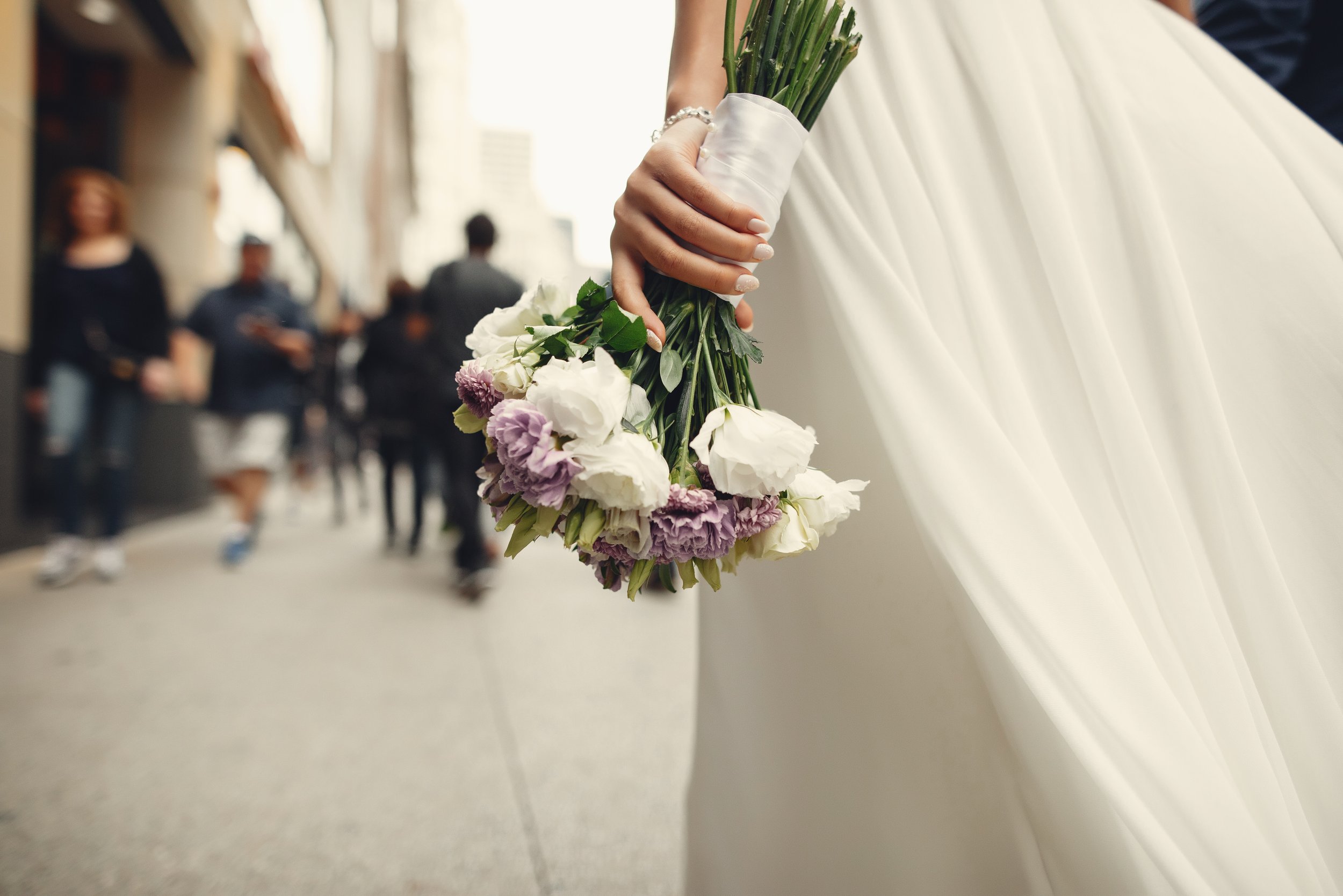Una mano con guantes blancos sostiene un ramo de flores blancas y moradas, en un vestido blanco de boda en una calle concurrida.