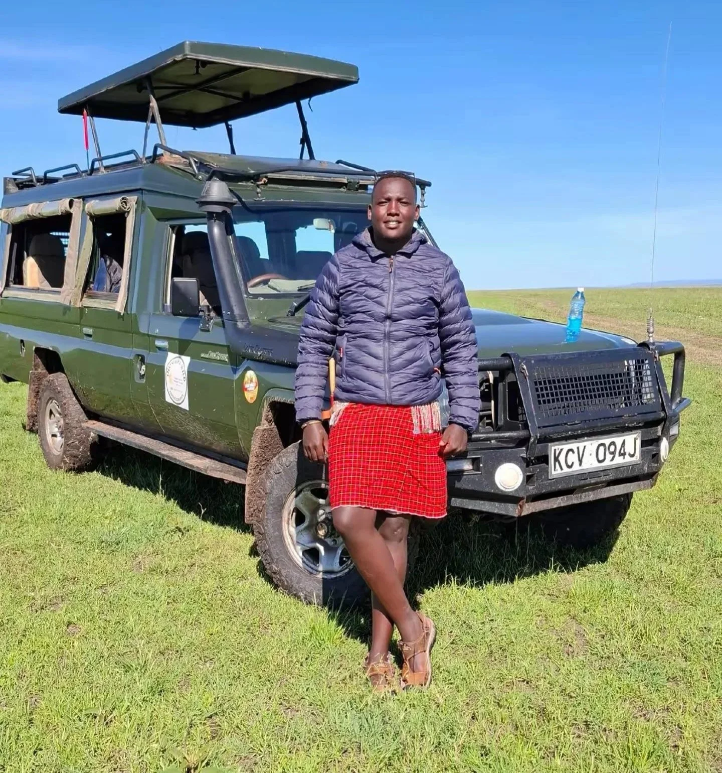 A man standing outdoors next to a green safari vehicle under a clear blue sky.