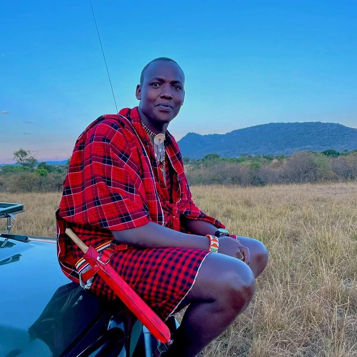 A man dressed in traditional Maasai clothing, sitting on a black vehicle in a grassy plain with hills in the background during sunset.