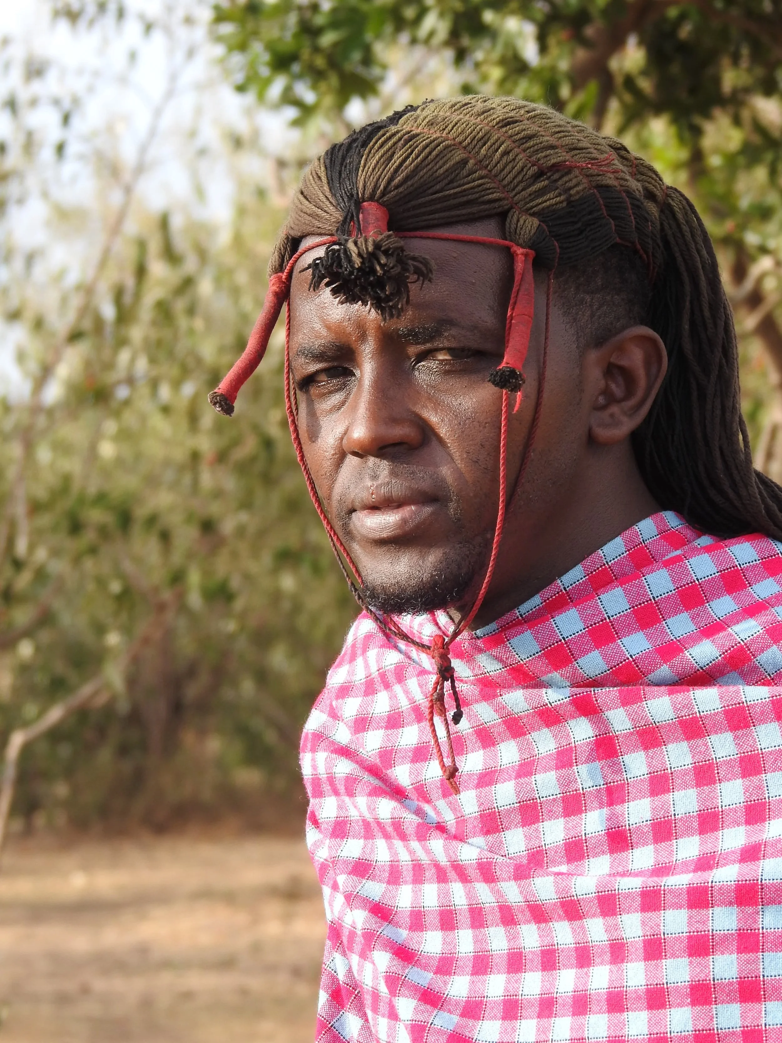 A man with medium dark skin, wearing a checkered pink and white shirt, has dreadlocks and a red headband or headpiece, outdoors with trees and blurred background.