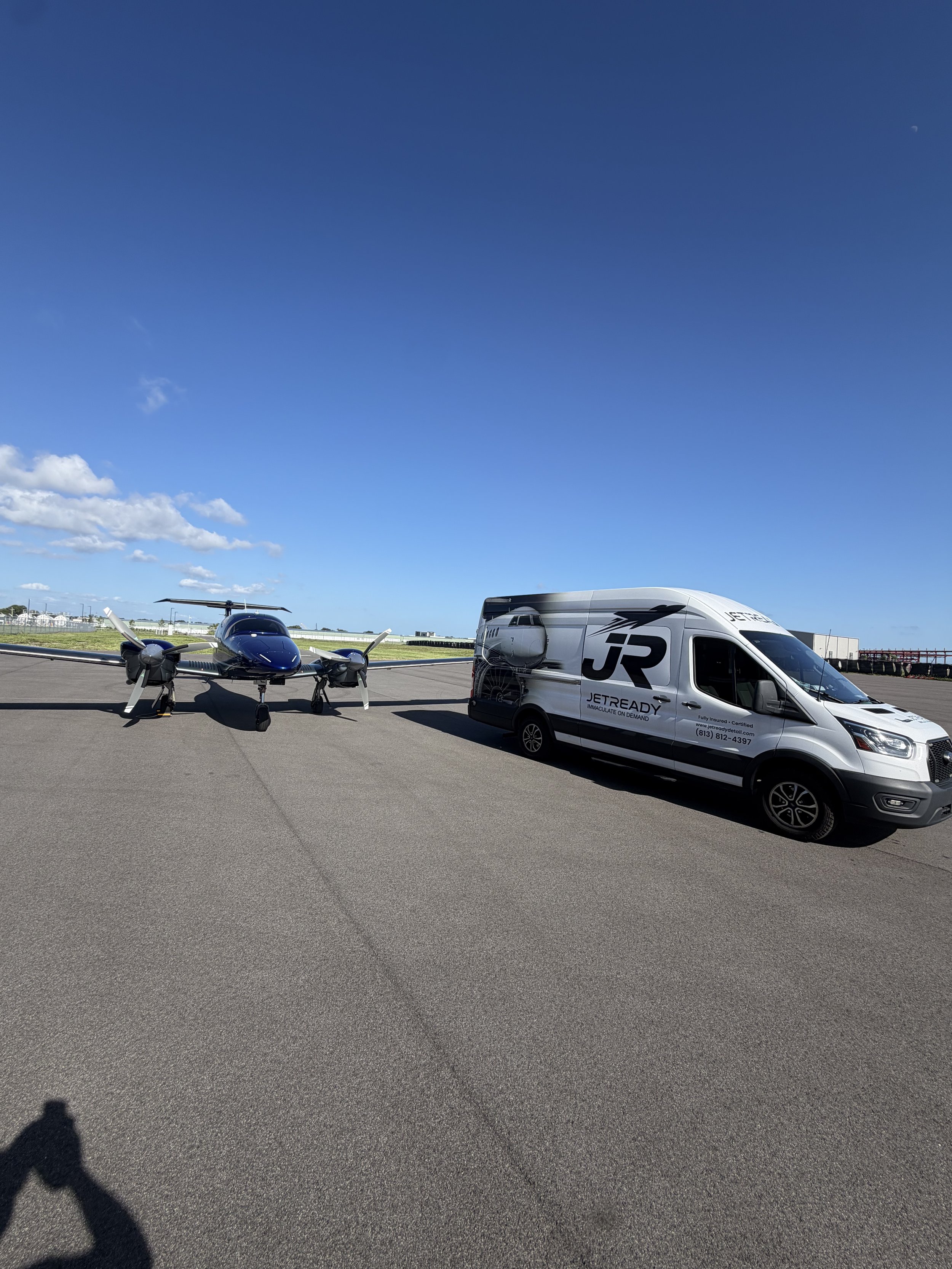 A small aircraft, a van with branding, and part of a shadow of a person taking the photo on an airport tarmac under a blue sky.