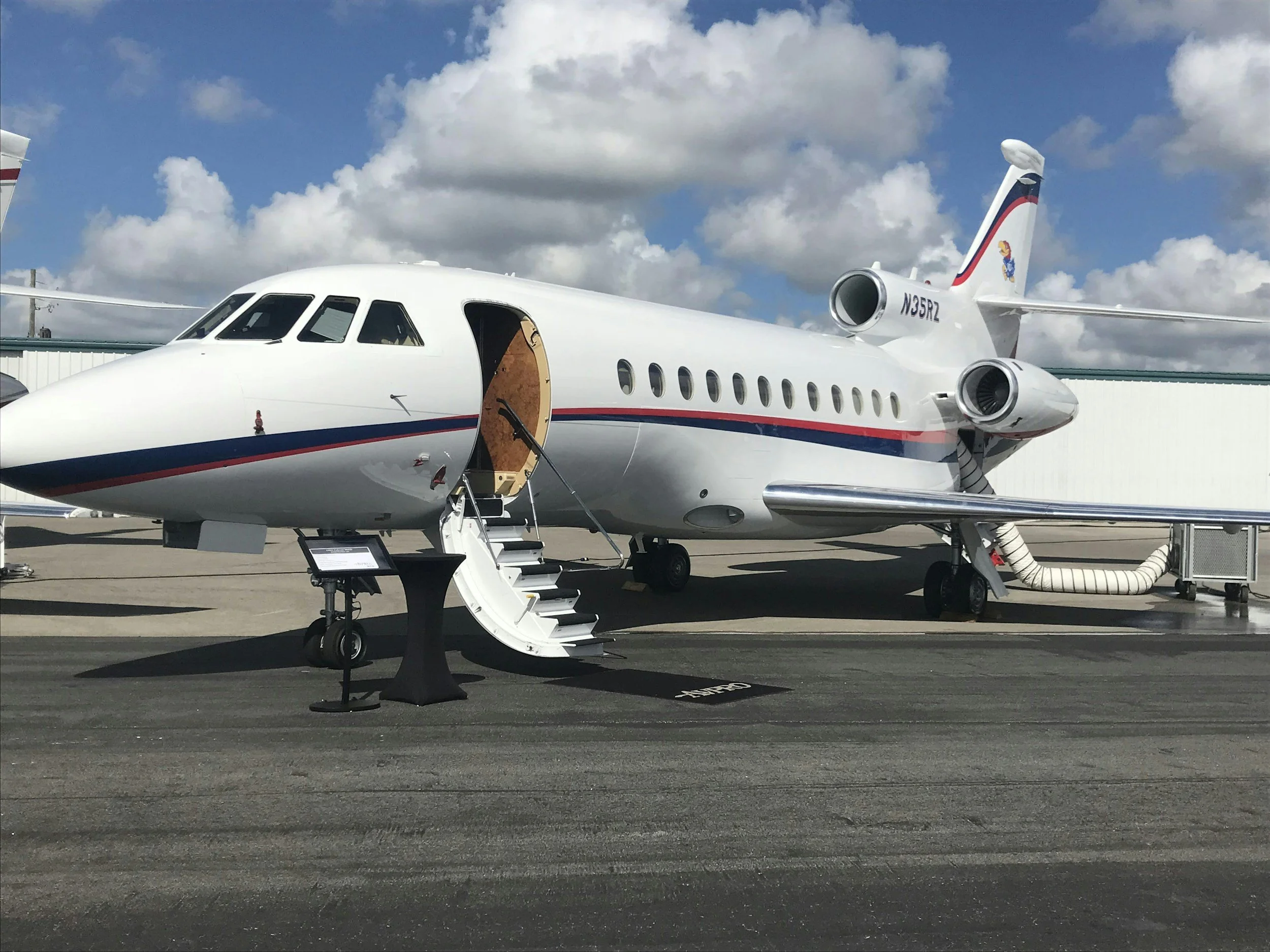 A white private jet with a red and blue stripe, parked on the tarmac with its door open and small stairs leading up to it, under a partly cloudy sky.