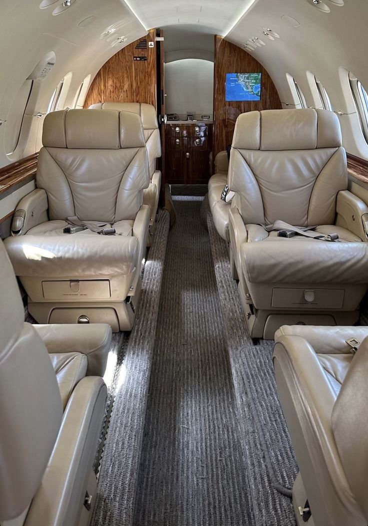Interior of a private jet with four beige leather seats, a carpeted aisle, and a wooden panel in the back with a TV screen showing a map.
