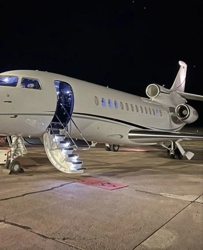 Private jet airplane parked on tarmac at night, with illuminated stairs leading into the aircraft.