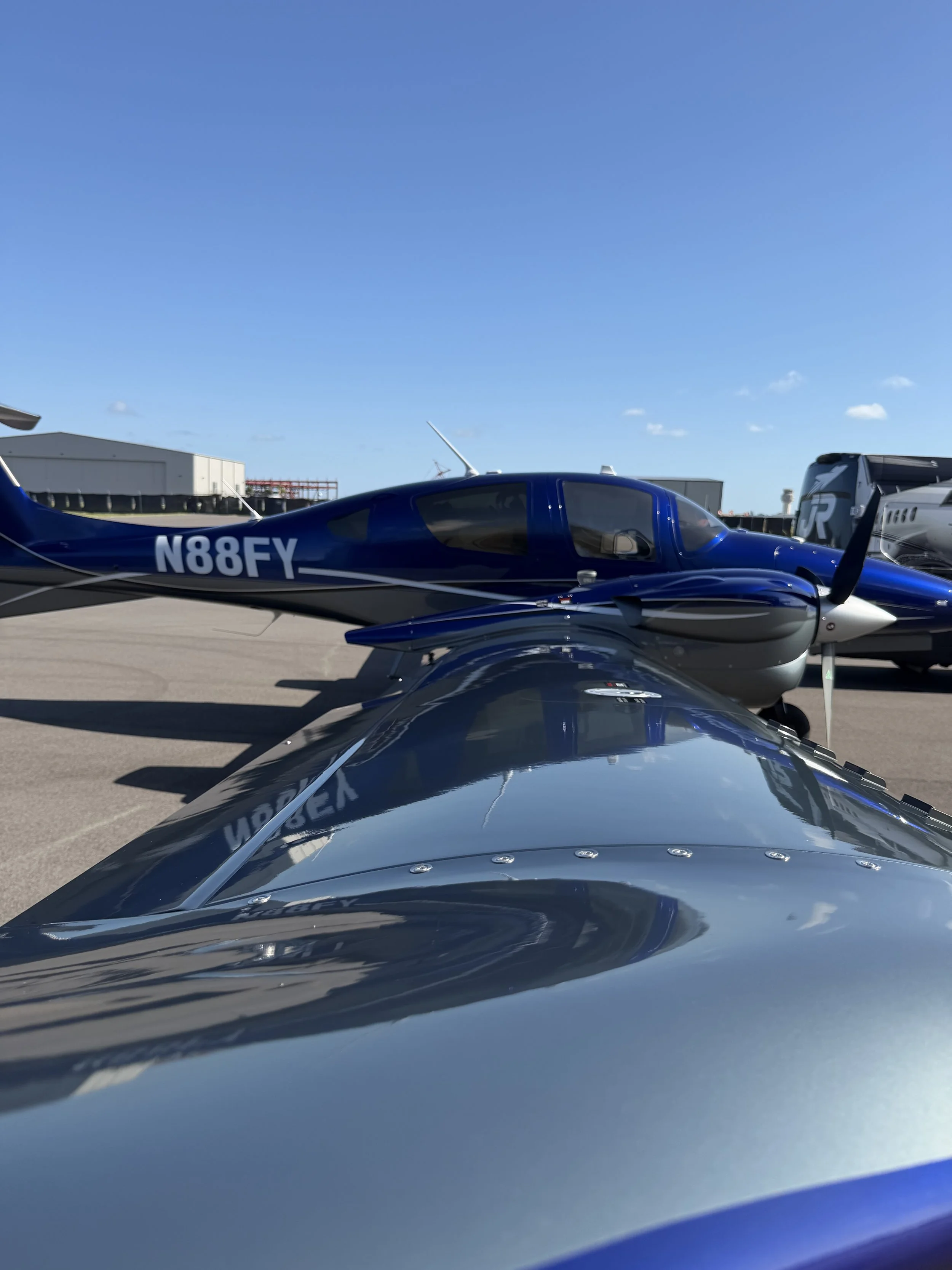 View of two aircraft parked on the tarmac at an airport under a clear blue sky, with hangars and airport facilities in the background.