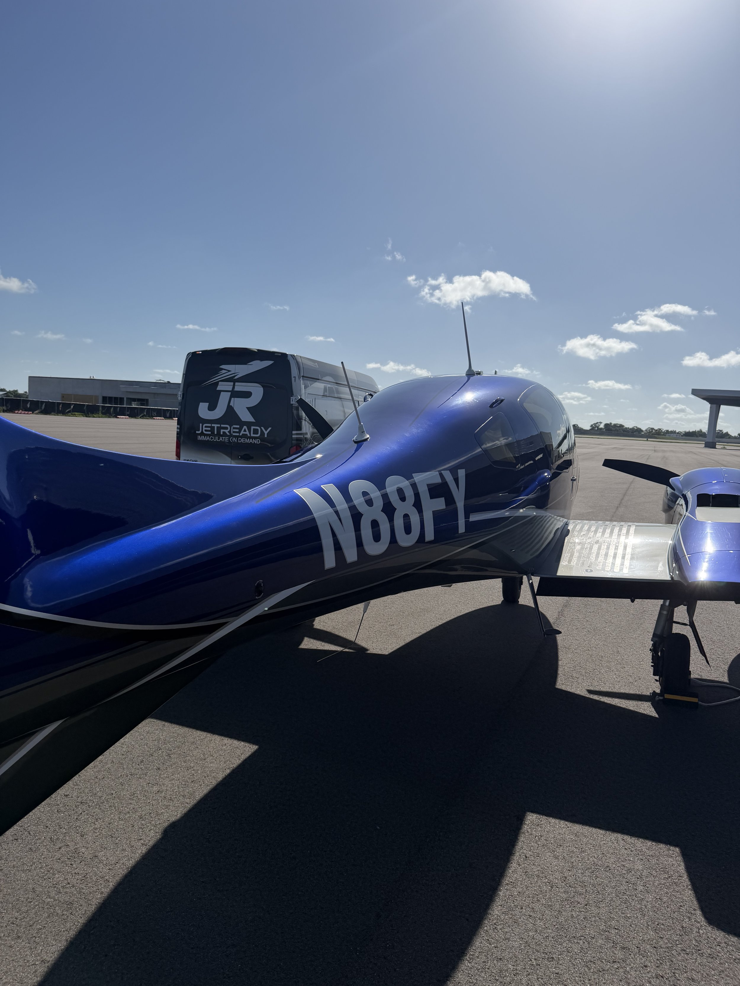 Blue racing airplane parked on the tarmac with a jet ready for takeoff in the background under a clear blue sky.