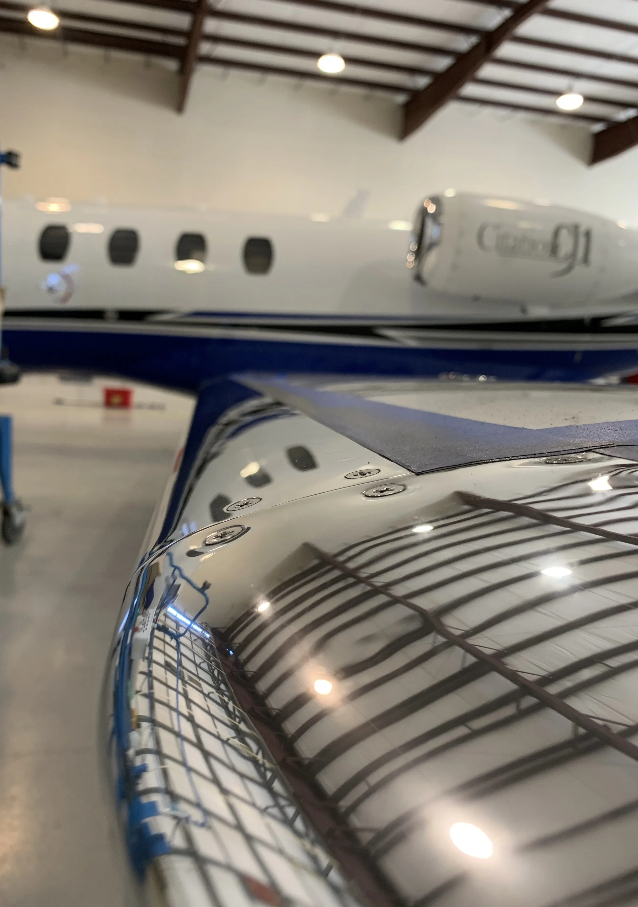 Close-up of the wings and tail of an airplane inside a hangar, with reflections on nearby metallic surfaces.