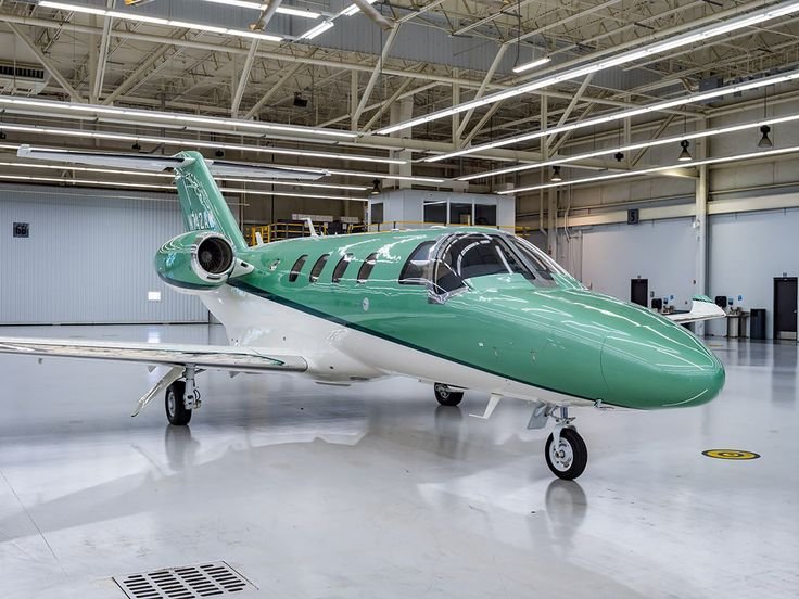 A green and white private jet aircraft inside a spacious hangar with white floors and ceiling, and some equipment visible in the background.