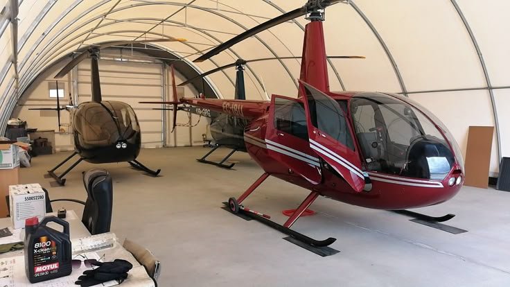 Red and black helicopters inside a spacious hangar with tools and chairs nearby.