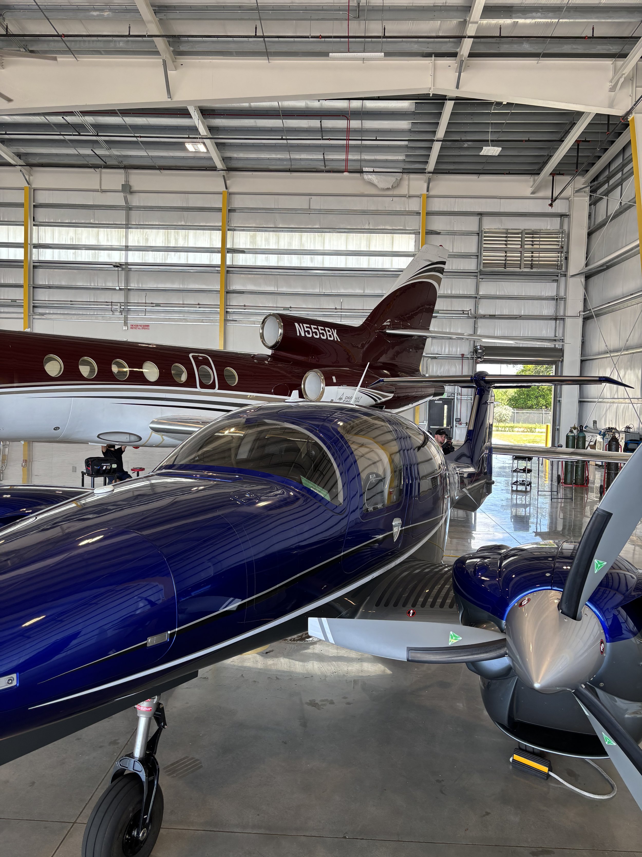View inside a hangar showing a blue and black aircraft and a maroon aircraft with a tail number N555BK, with various tools and equipment in the background.