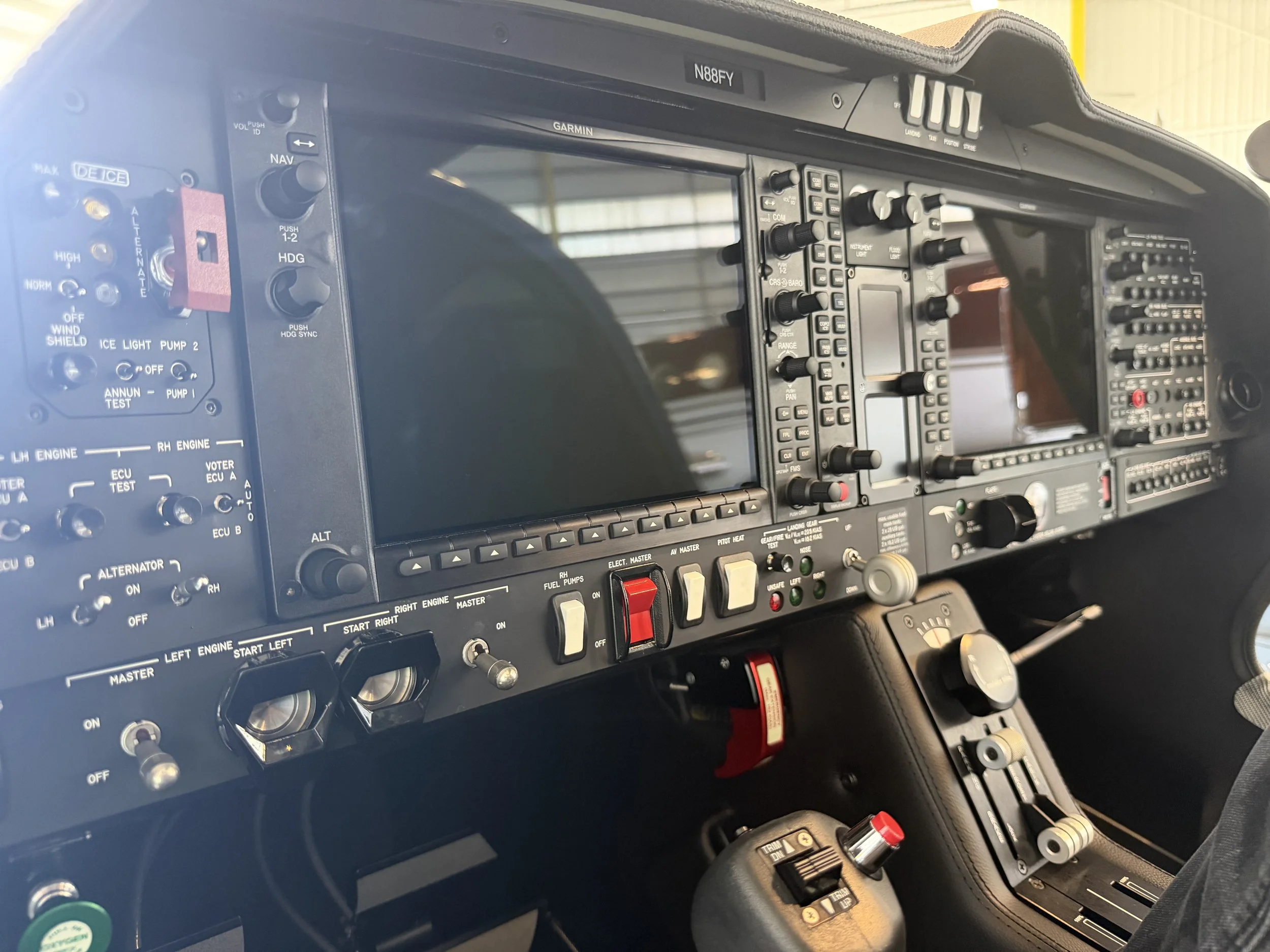 Close-up of an aircraft cockpit dashboard with various controls, switches, and gauges, including a Garmin display screen and labeled buttons.