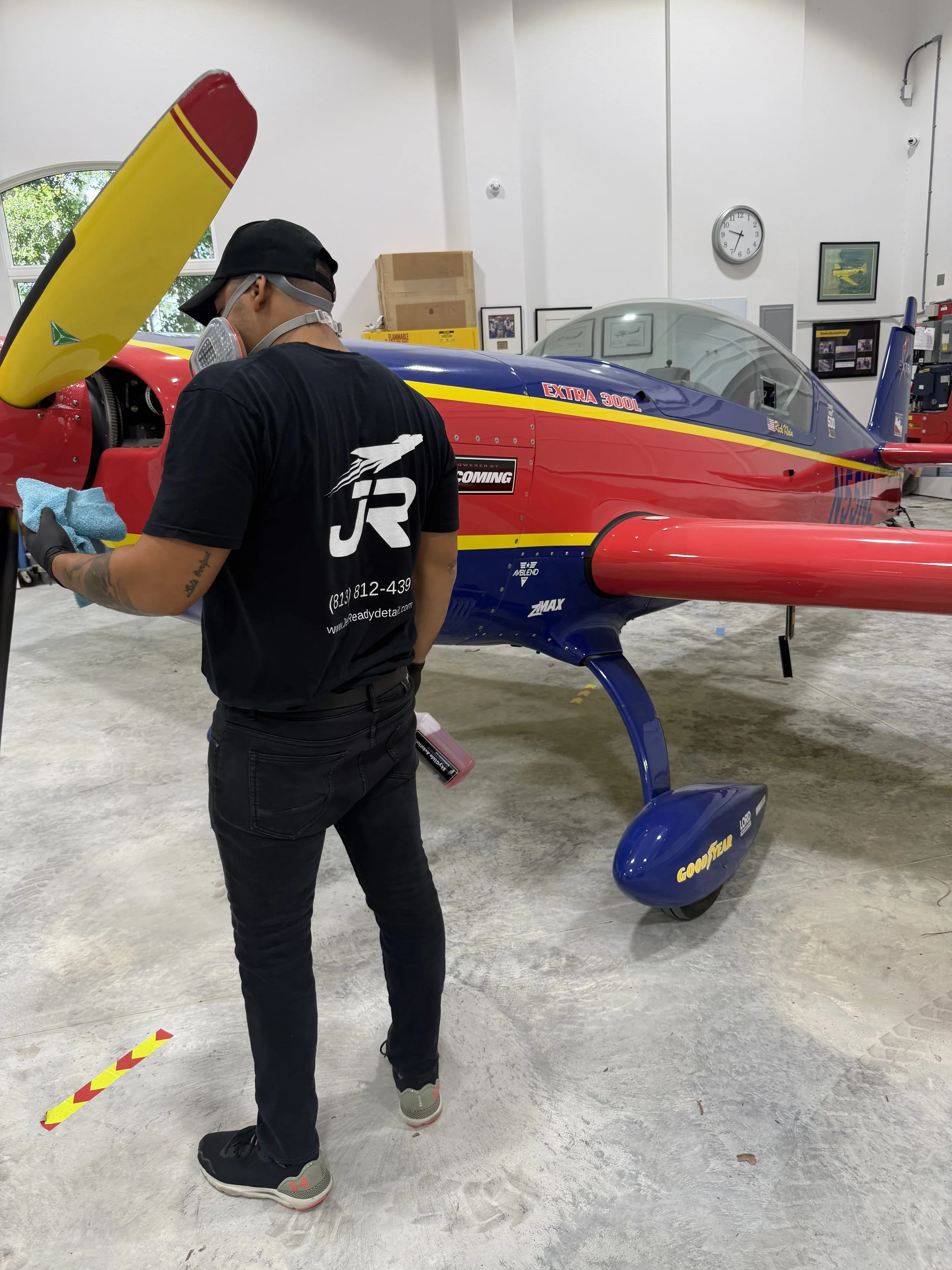 Technician wearing a black cap, face mask, and gloves working on a blue and red airplane inside a hangar. The airplane has a yellow stripe and the words 'Extra 300L' on the side. The technician holds a blue cloth while inspecting the plane.