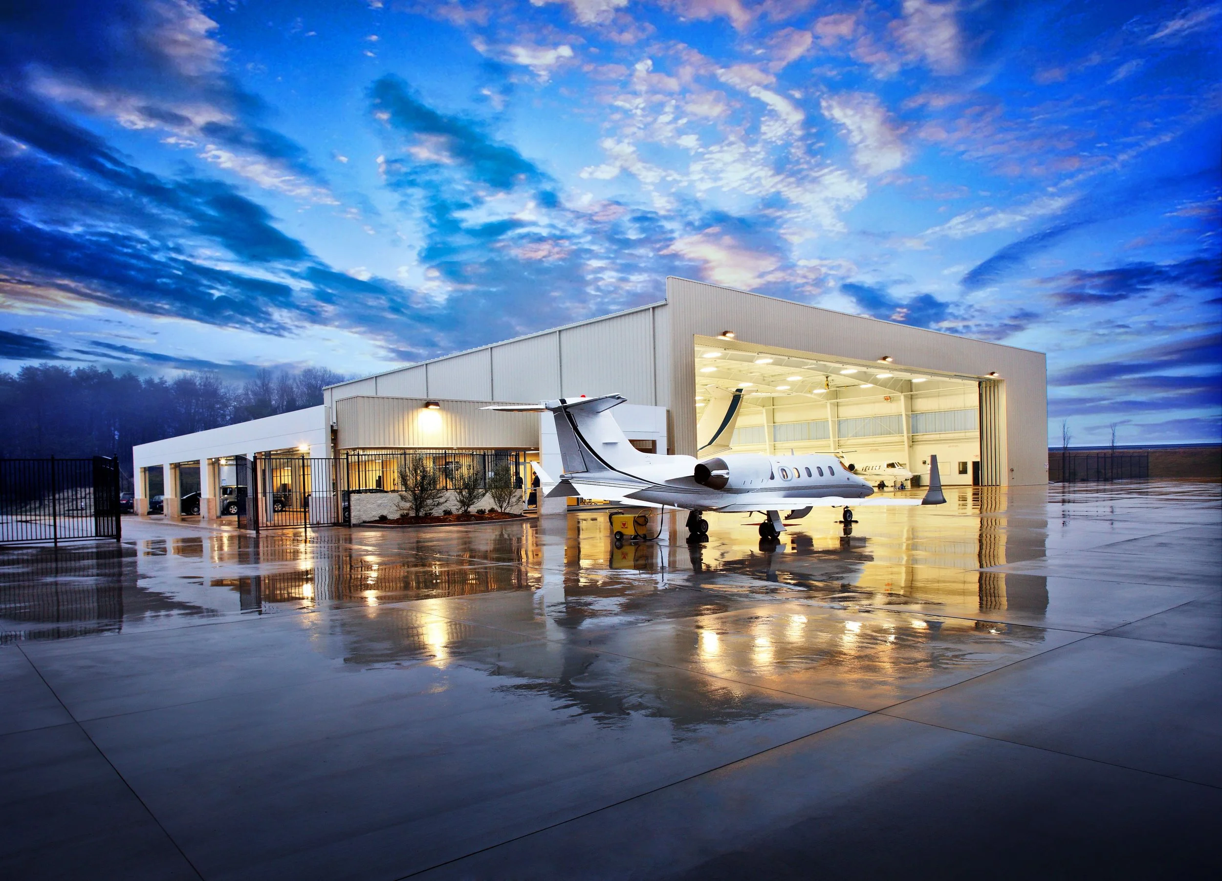 Private jet parked outside hangar at airport during dawn or dusk, with a dramatic blue and purple sky and reflective wet tarmac.