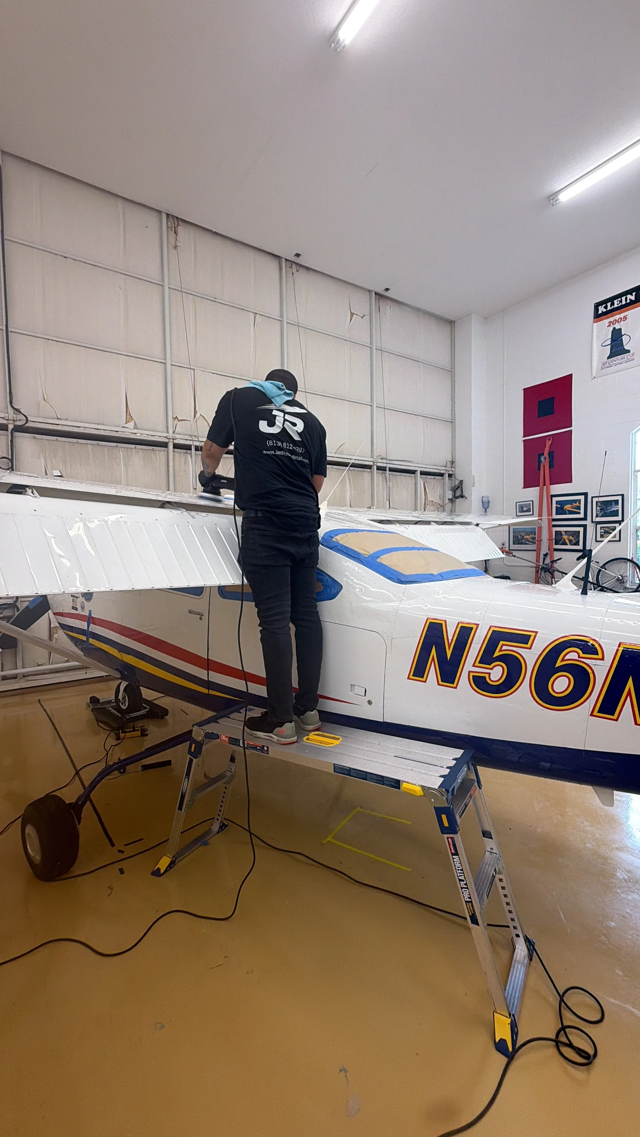 Person working on small airplane inside hangar, standing on ladder, wearing black shirt, with tools and equipment around.