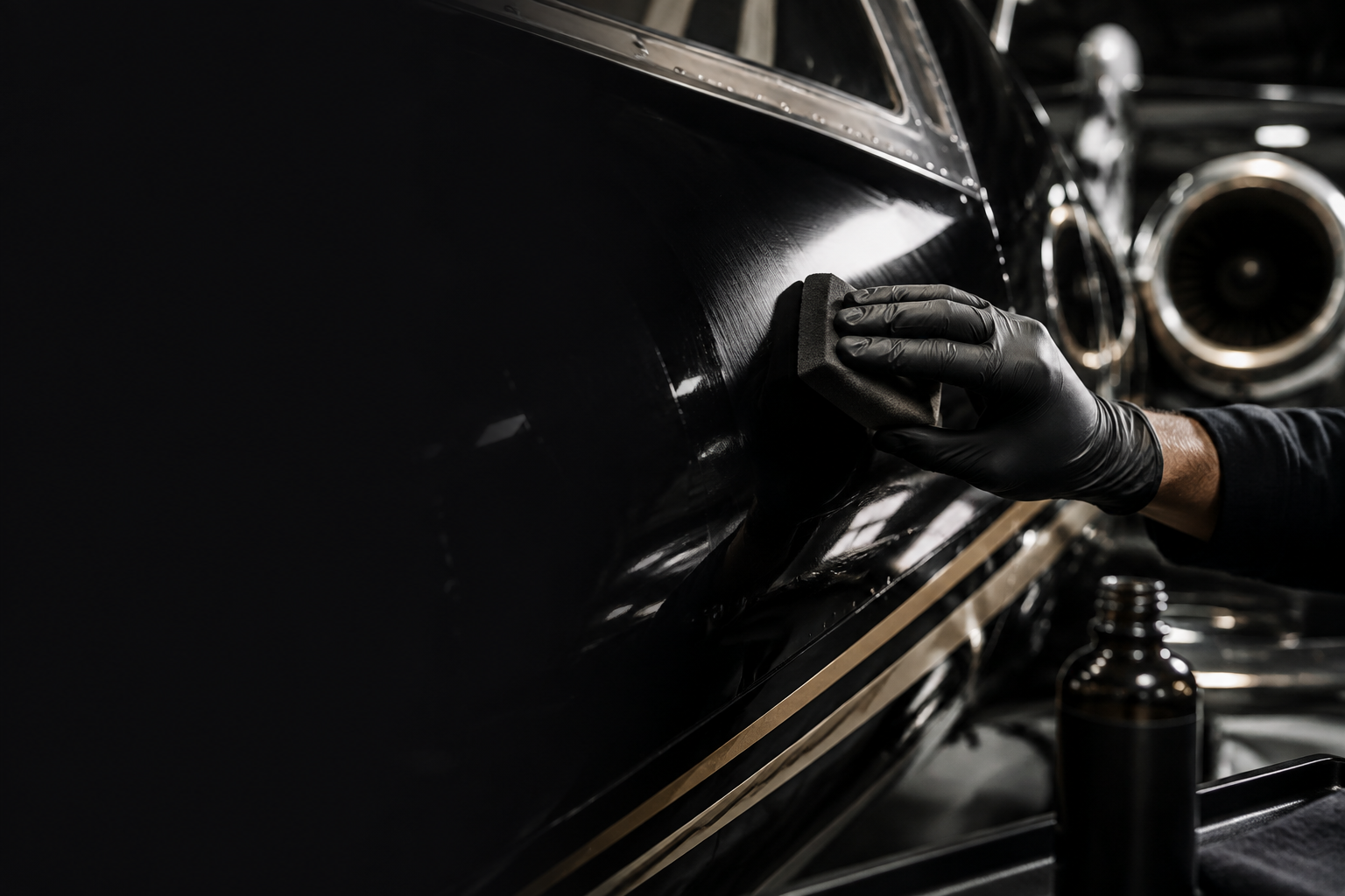 Person wearing black gloves polishing a black vintage car with a cloth in a dimly lit garage.