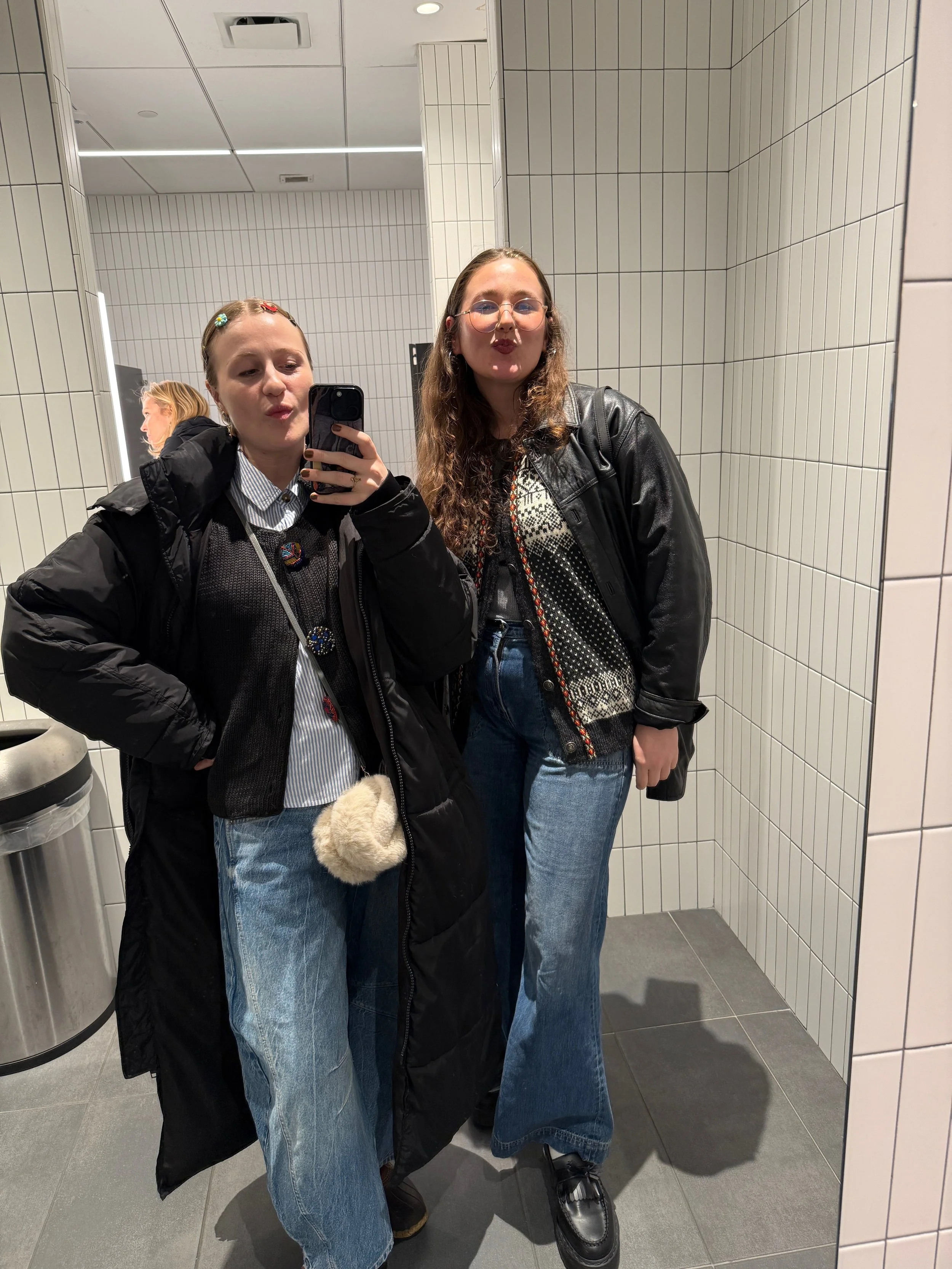 Two young women taking a mirror selfie in a bathroom with white tiled walls and gray floor tiles. They are both wearing black jackets and blue jeans, with one woman making a kissy face and the other pursing her lips.