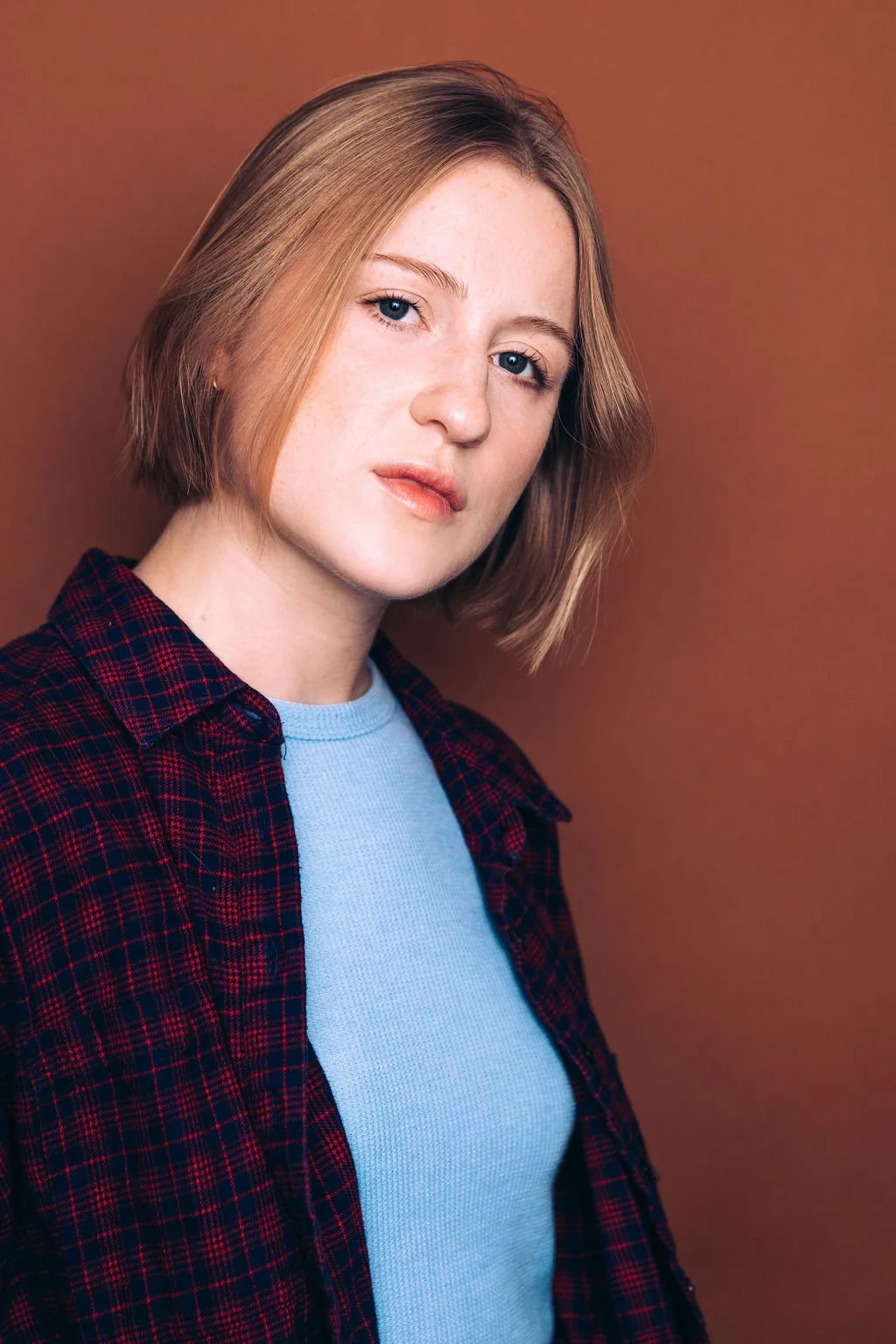 Portrait of a young woman with short blonde hair, blue eyes, wearing a light blue shirt and red plaid jacket, against a reddish-brown background.