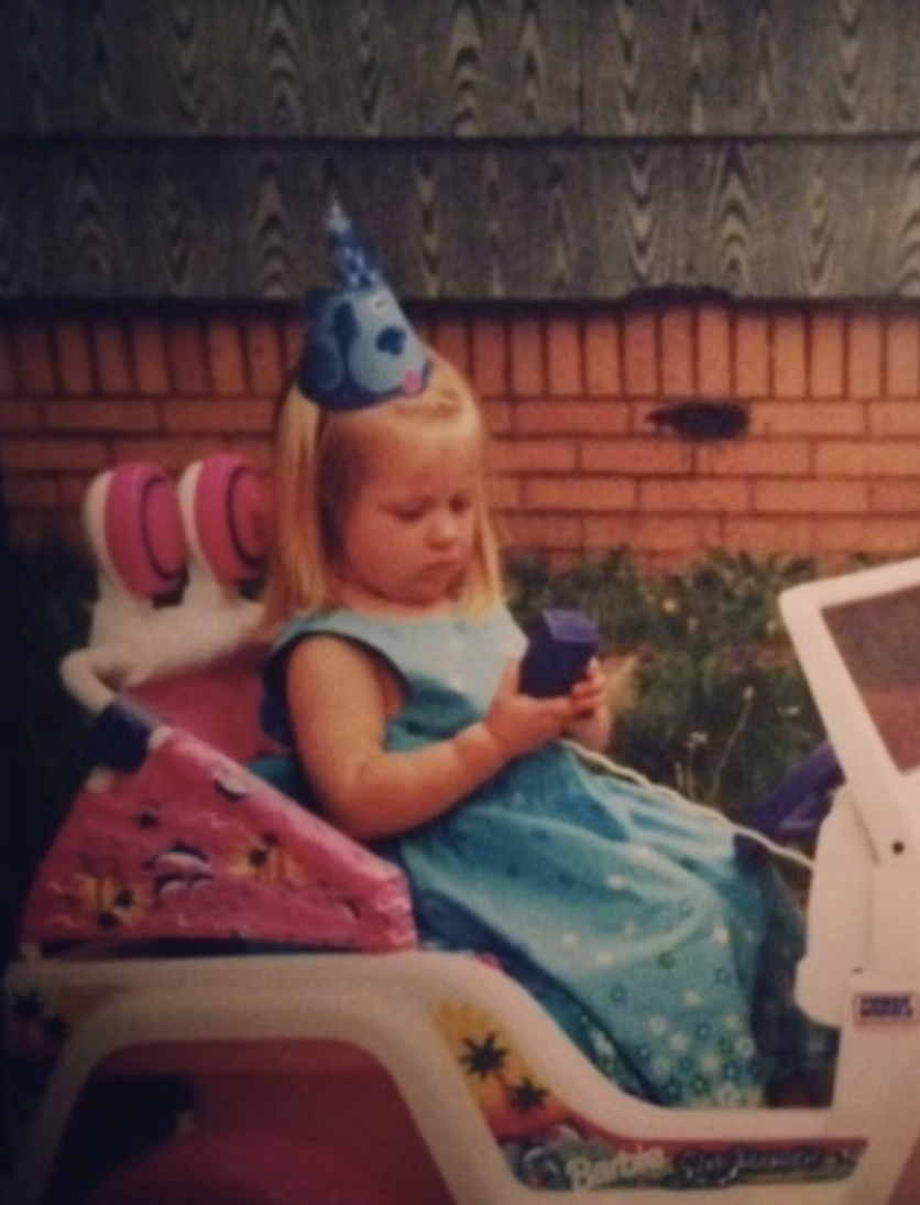 Young girl sitting outdoors in a pink kids car, wearing a birthday party hat and a blue dress, looking at a device in her hands.