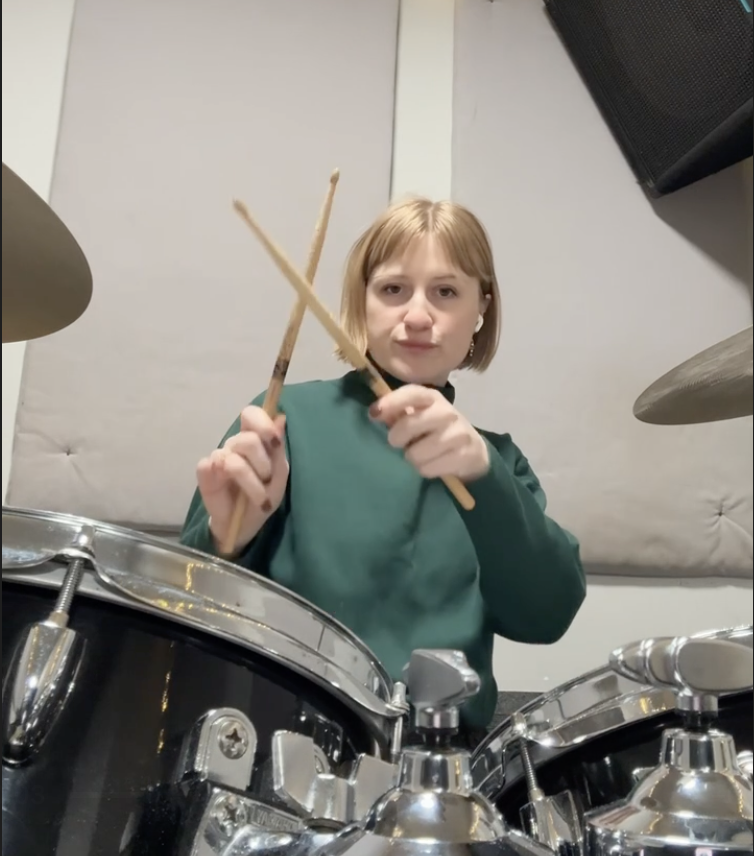 Young woman playing drums in a music studio, holding drumsticks and looking at the camera.