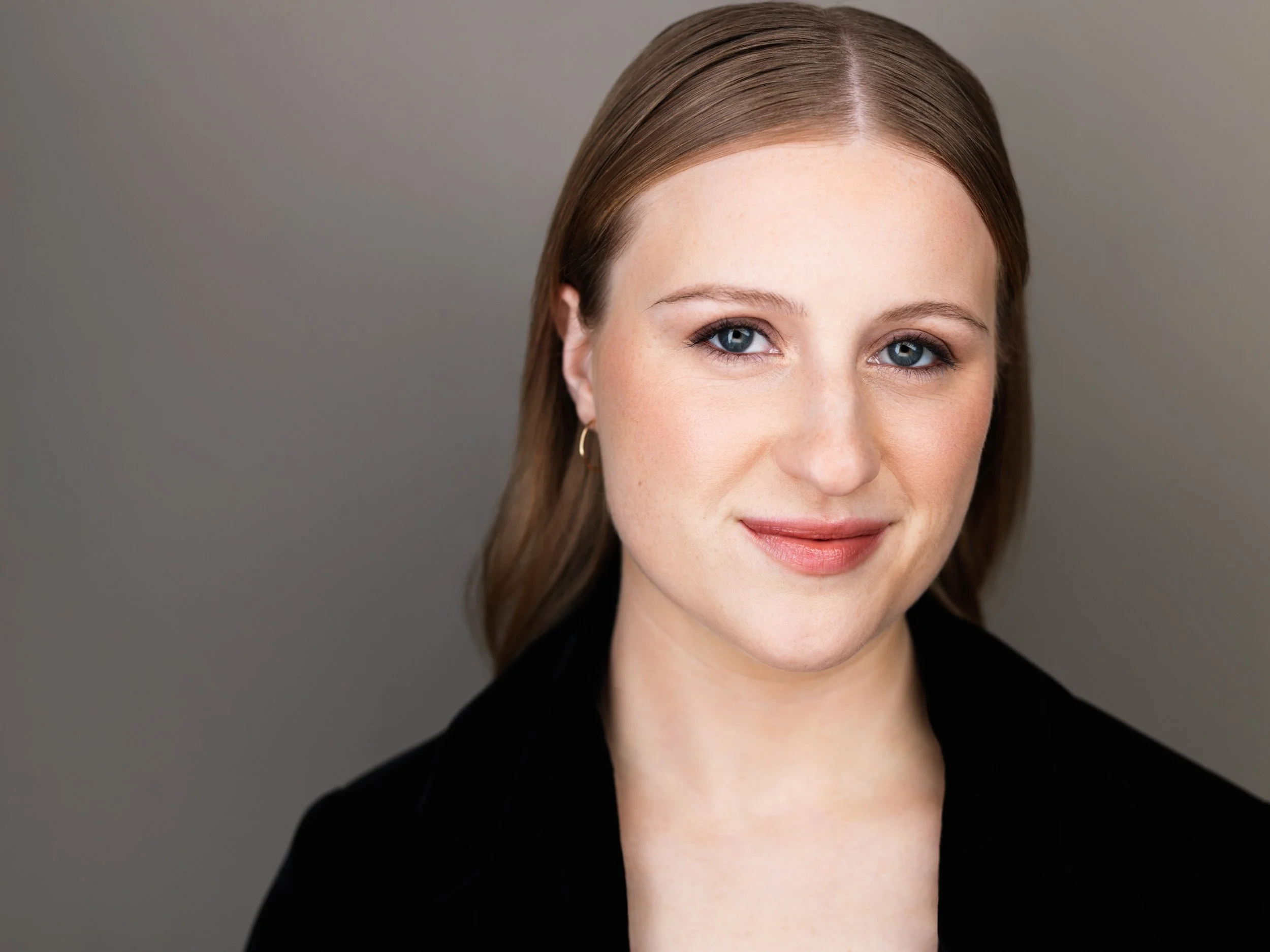 A woman with fair skin, reddish blonde hair, and blue eyes, wearing gold earrings and a black top, smiling softly against a gray background.