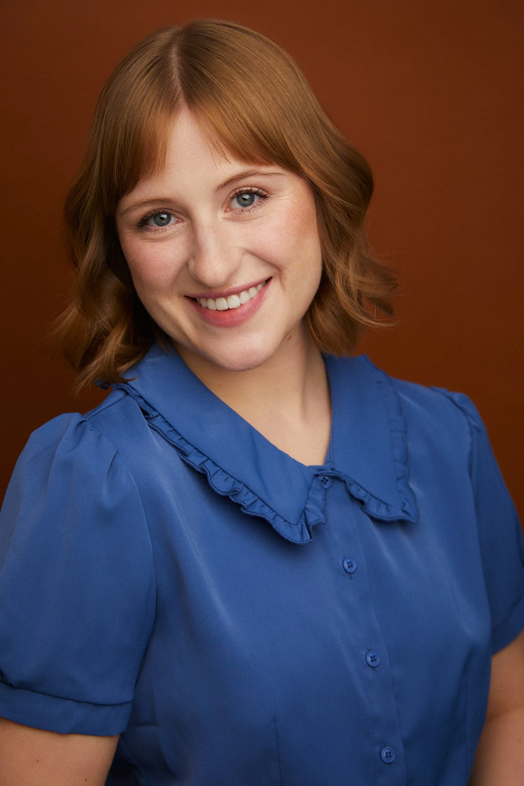Portrait of a young woman with reddish blonde hair, blue eyes, and a warm smile, wearing a blue blouse with ruffled collar, against a brown background.