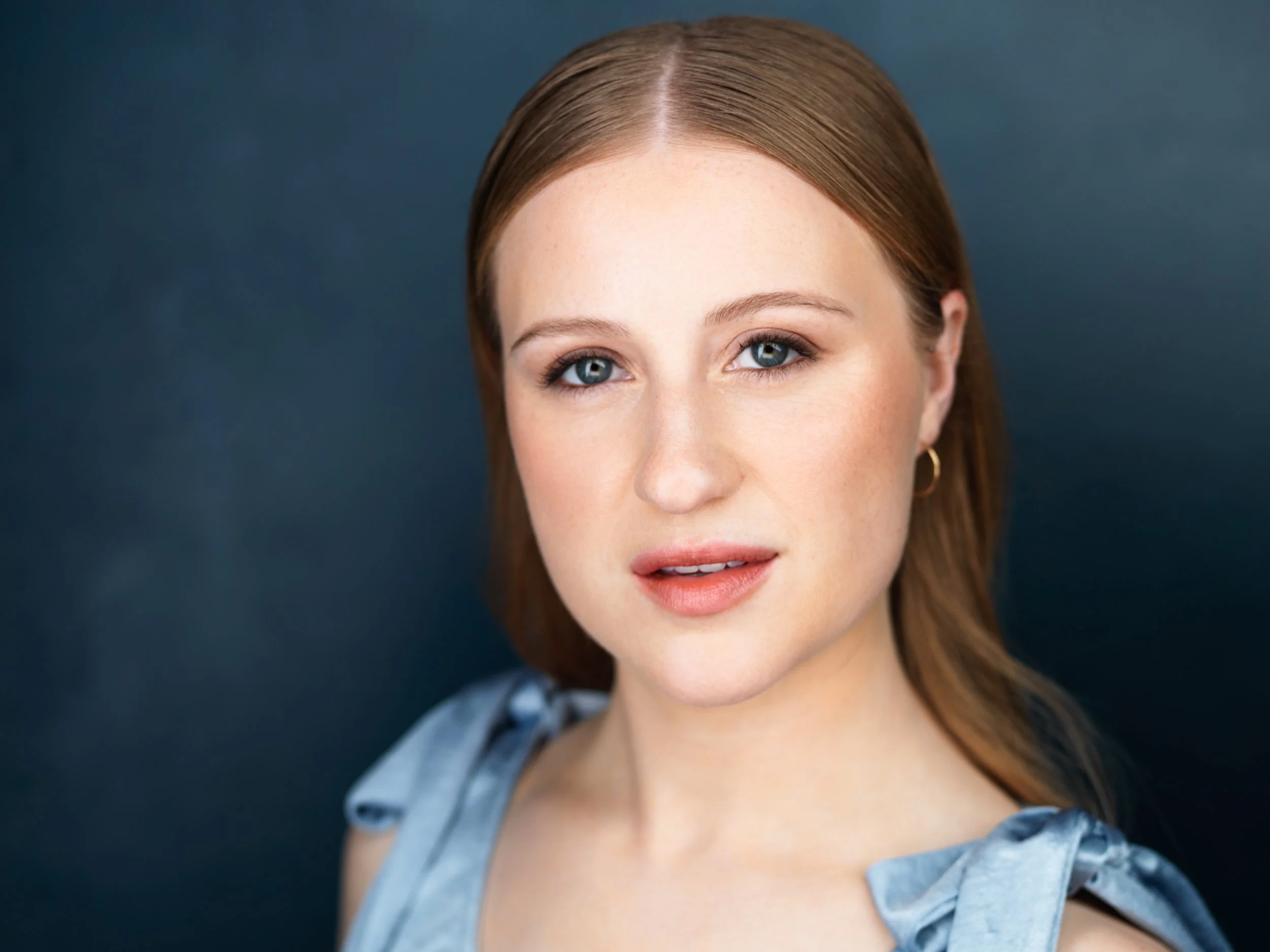 Close-up portrait of a woman with fair skin, blue eyes, and reddish-blonde hair, wearing a blue shirt with shoulder bows, gold hoop earrings, and soft makeup, against a dark background.