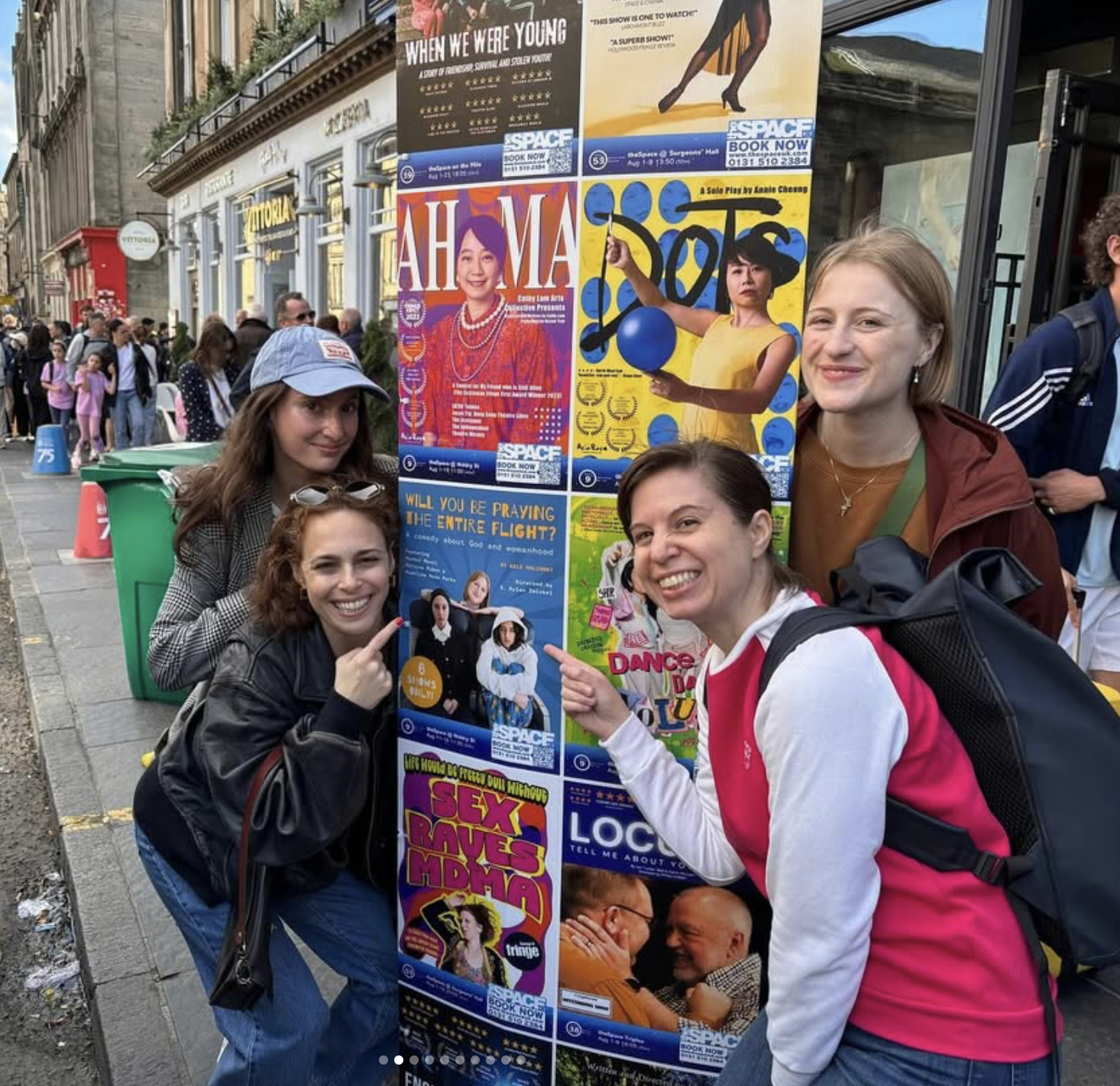 Four women smiling and pointing at a display board of theater posters on a city sidewalk. The posters advertise various plays, including 'Ah Ma' and others. People are walking in the background.