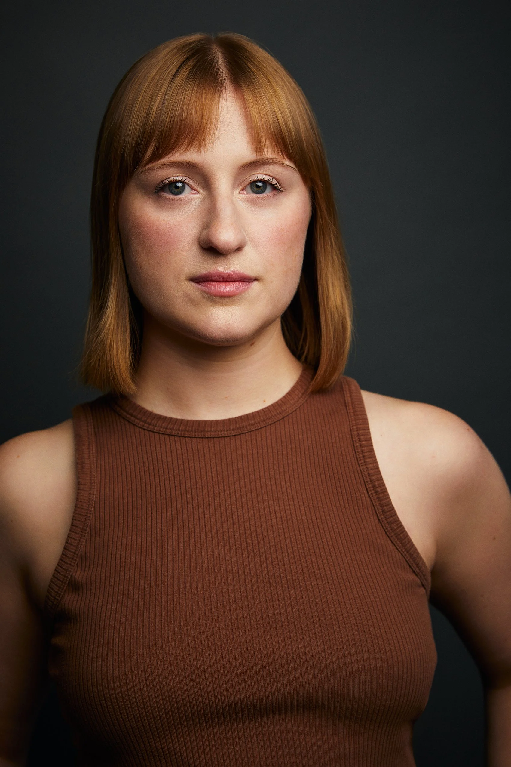 Portrait of a young woman with straight, reddish blonde hair, wearing a sleeveless, ribbed, brown top, against a dark background.