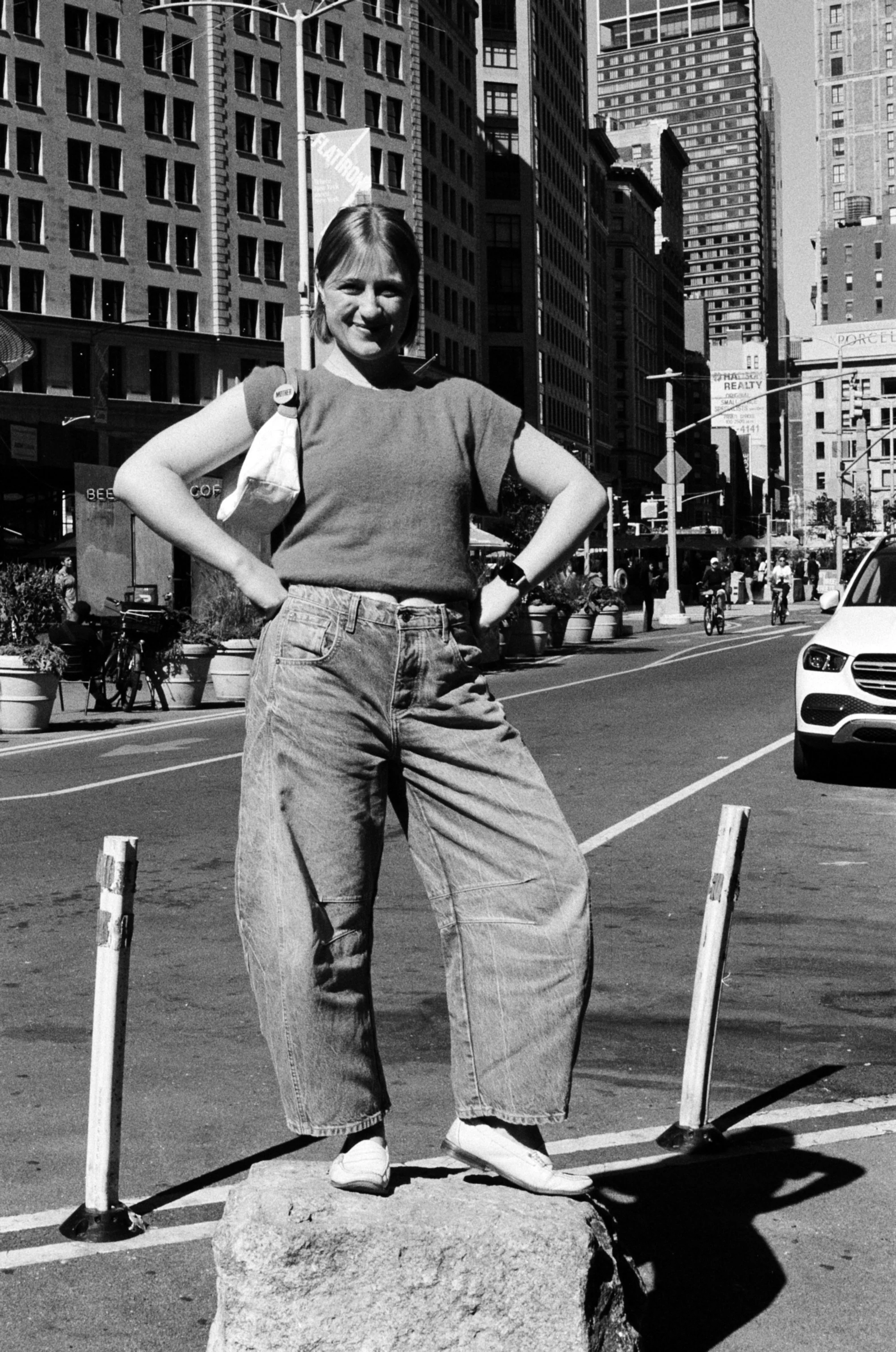 A woman standing on a large rock in the middle of a city street, posing with hands on hips. She is wearing barrel jeans, a short-sleeved top, and white loafers. The background features tall buildings, bicycles, and cars.