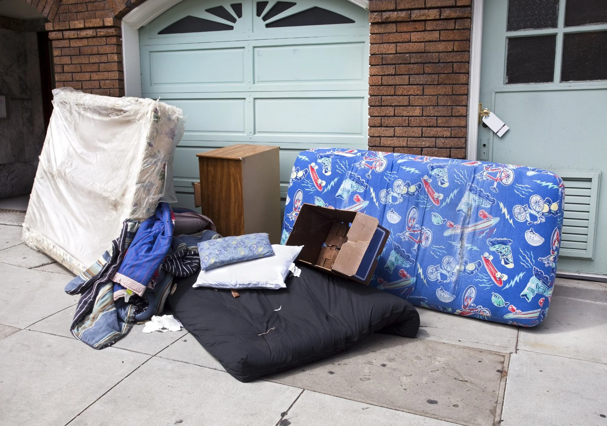 Street scene with discarded household furniture and items in front of a garage. Items include a mattress, pillows, a wooden nightstand, two mattresses, clothes, and a cardboard box, all lying on the sidewalk.