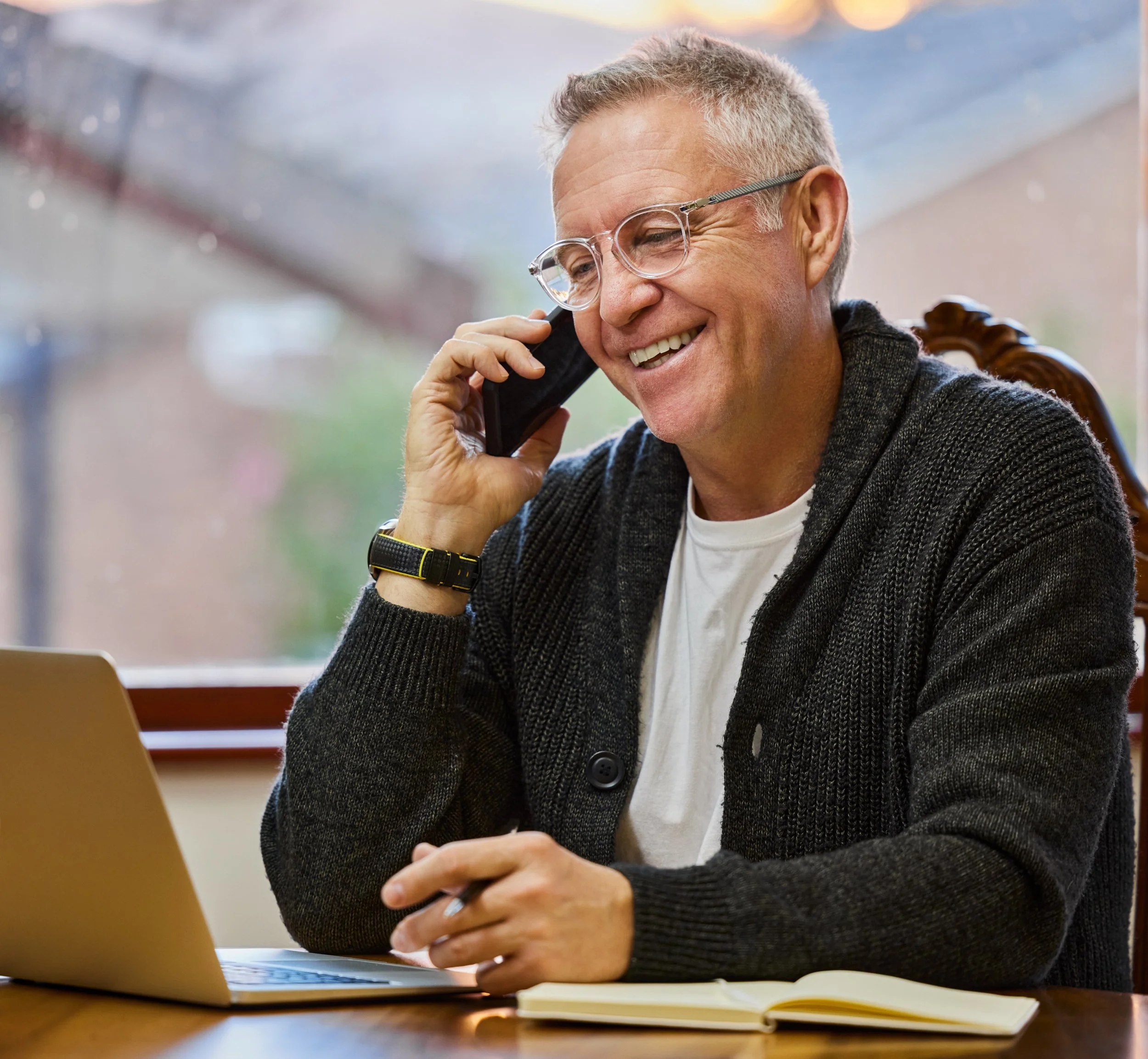Smiling older man with glasses talking on a cell phone while sitting at a desk with a laptop and notebook.