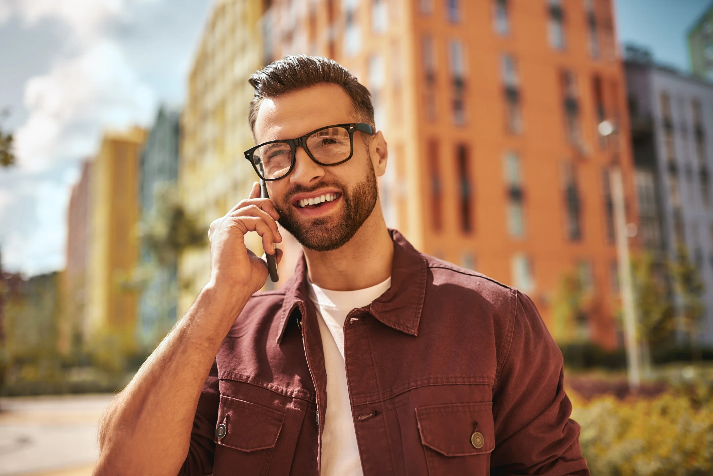Young man in glasses talking on a cell phone outdoors with colorful apartment buildings in the background.