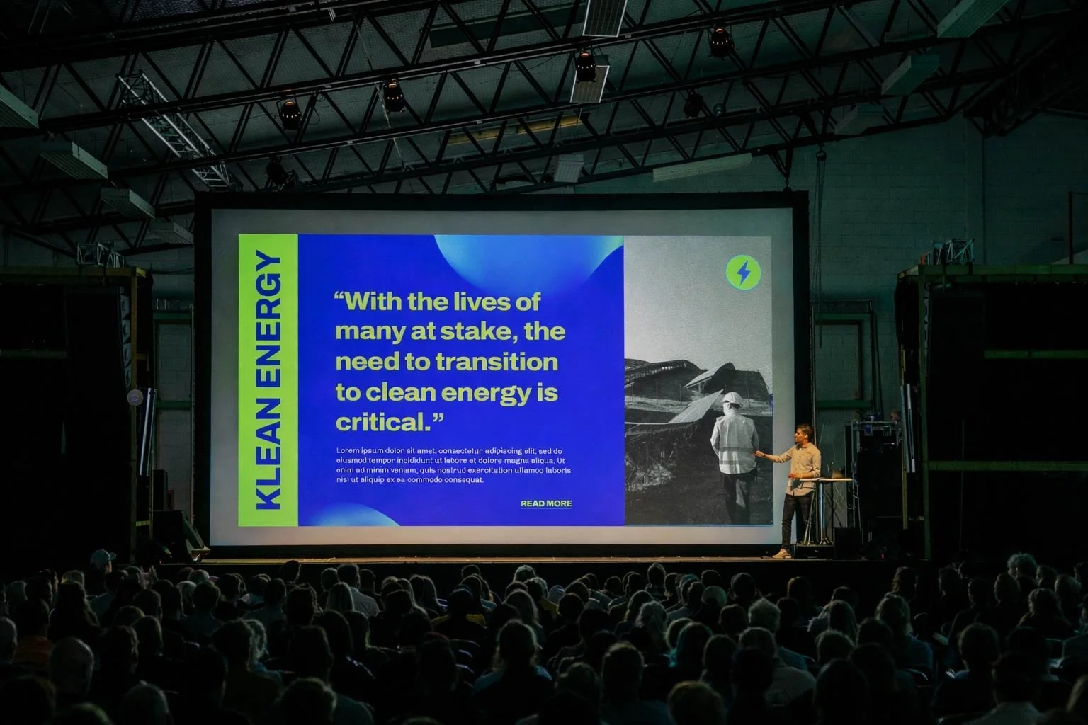 A presentation at a conference on clean energy, with a large screen displaying a quote about transitioning to clean energy, an image of wind turbines, and a speaker handing a mic to a participant on stage, amid a seated audience.