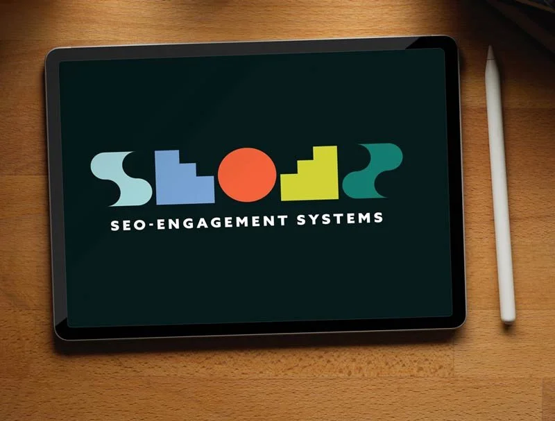 A tablet displaying a logo with colorful letters spelling 'SOLU' and 'SEO-ENGAGEMENT SYSTEMS' on a wooden desk next to a stylus pen.
