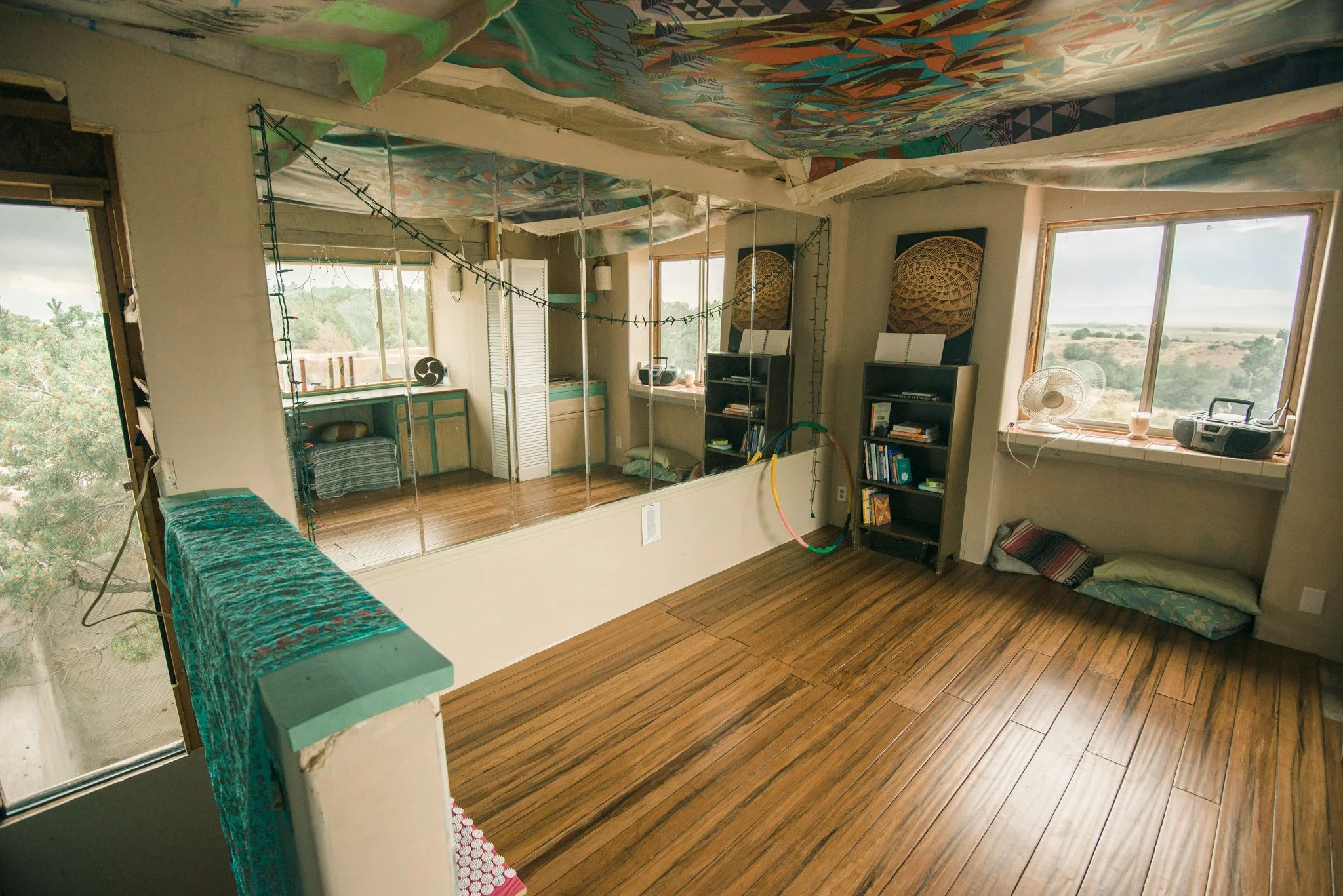 Living room with large windows, hardwood floors, bookshelf, fan, and colorful ceiling mural, partially under construction or renovation.