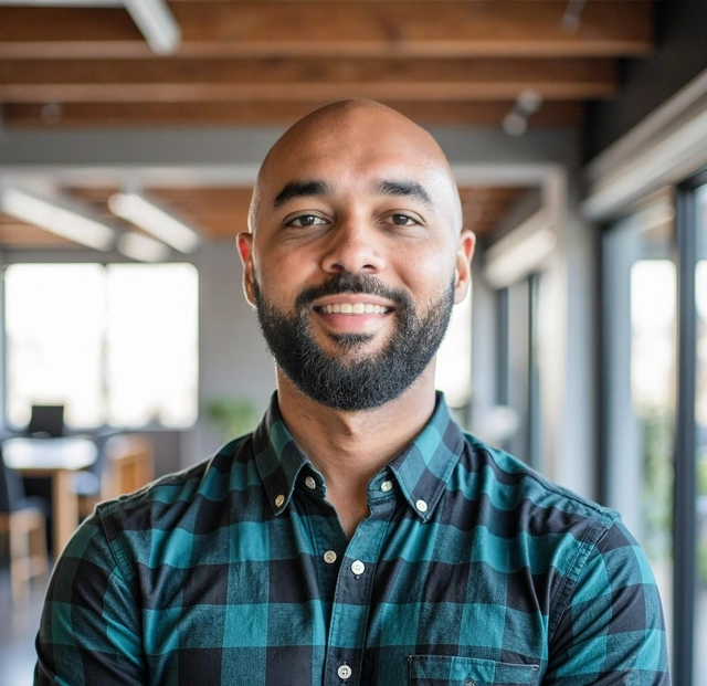 A smiling bald man with a beard wearing a green and blue checkered shirt in a modern office with large windows and wooden ceiling beams.