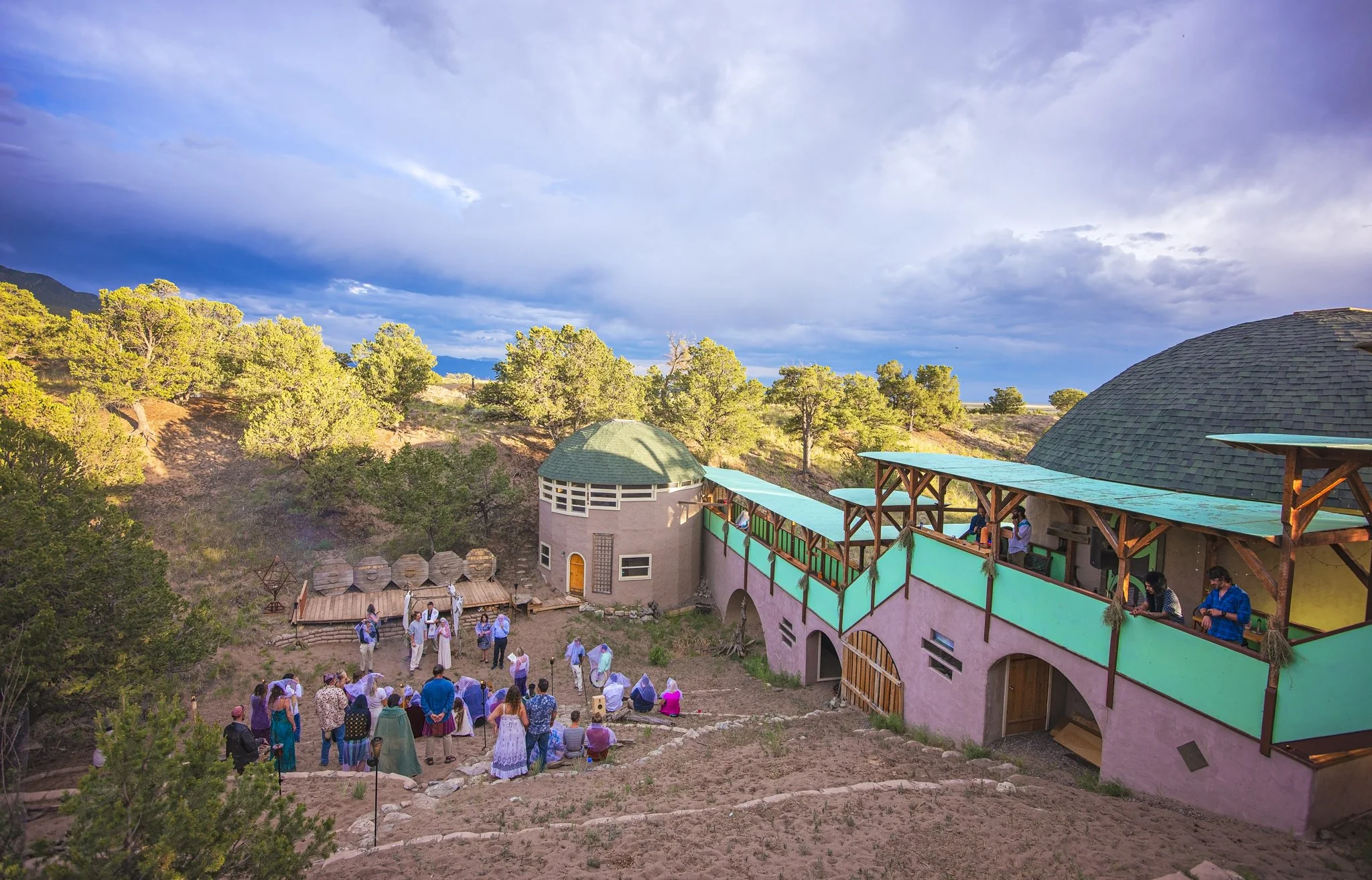 A group of people gathered outside a pink building with green roofing, surrounded by trees and hilly terrain under a partly cloudy sky.