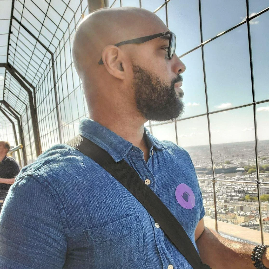 A man with sunglasses, a beard, and a shaved head looking out from a tall observation tower with a city view and a wire mesh fence.