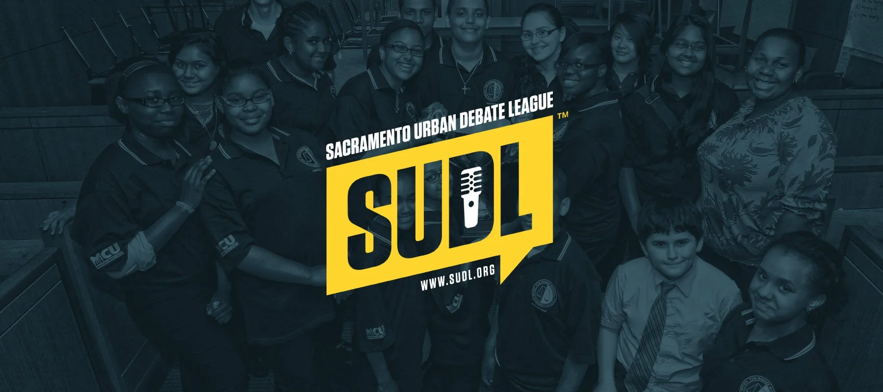Group of young students and a teacher in a classroom, holding a sign that reads 'Sacramento Urban Debate League.'