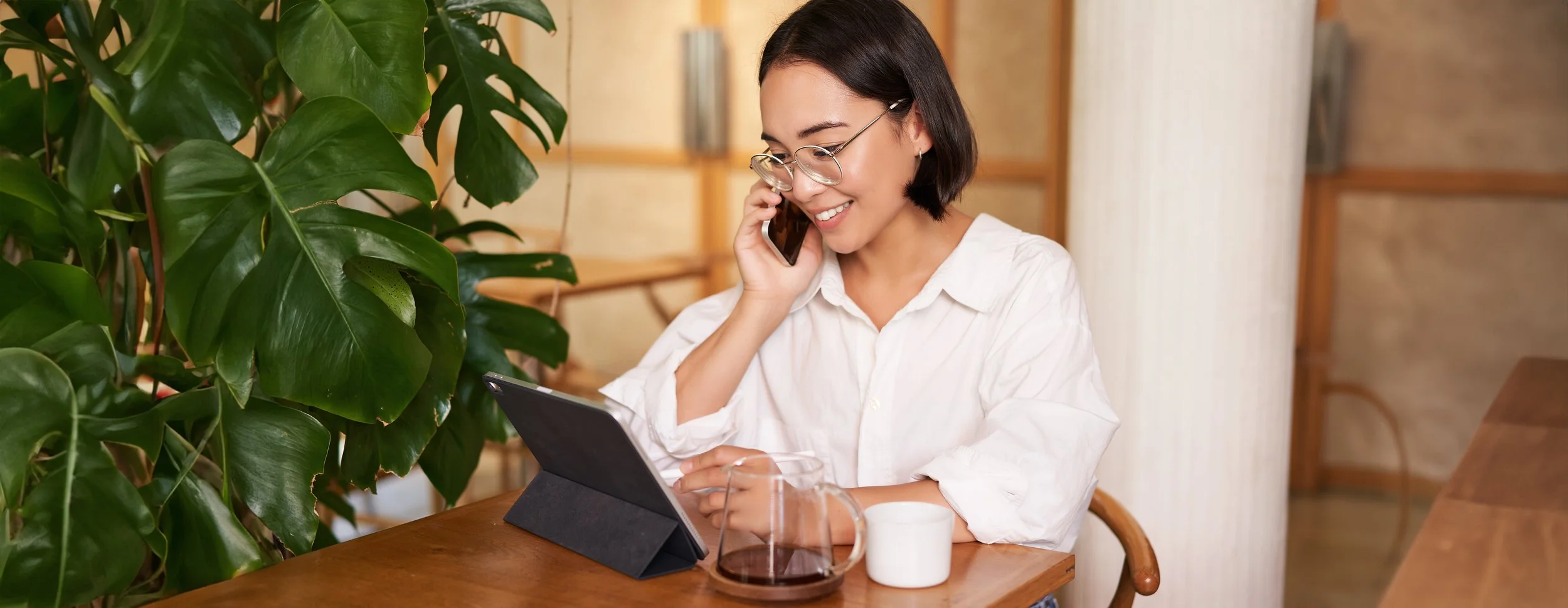A woman with short dark hair and glasses is smiling while talking on her cell phone. She is sitting at a wooden table with a tablet, a mug, and a glass carafe in front of her. Large green leafy plants are on her left, and a beige curtain and wooden shelves are in the background.
