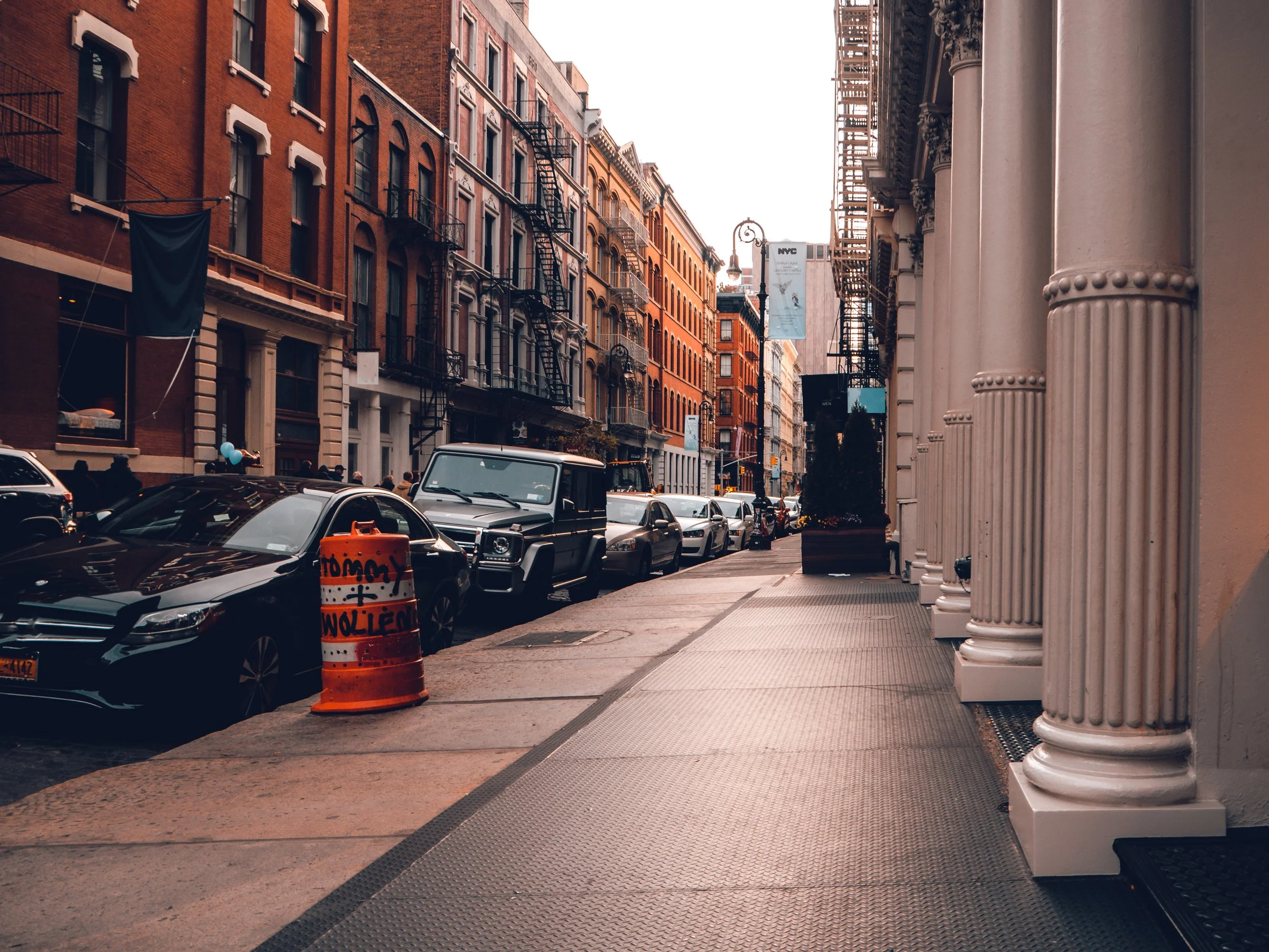 City street scene with parked cars, tall brick and stone buildings, fire escapes, and scaffolding on the right.