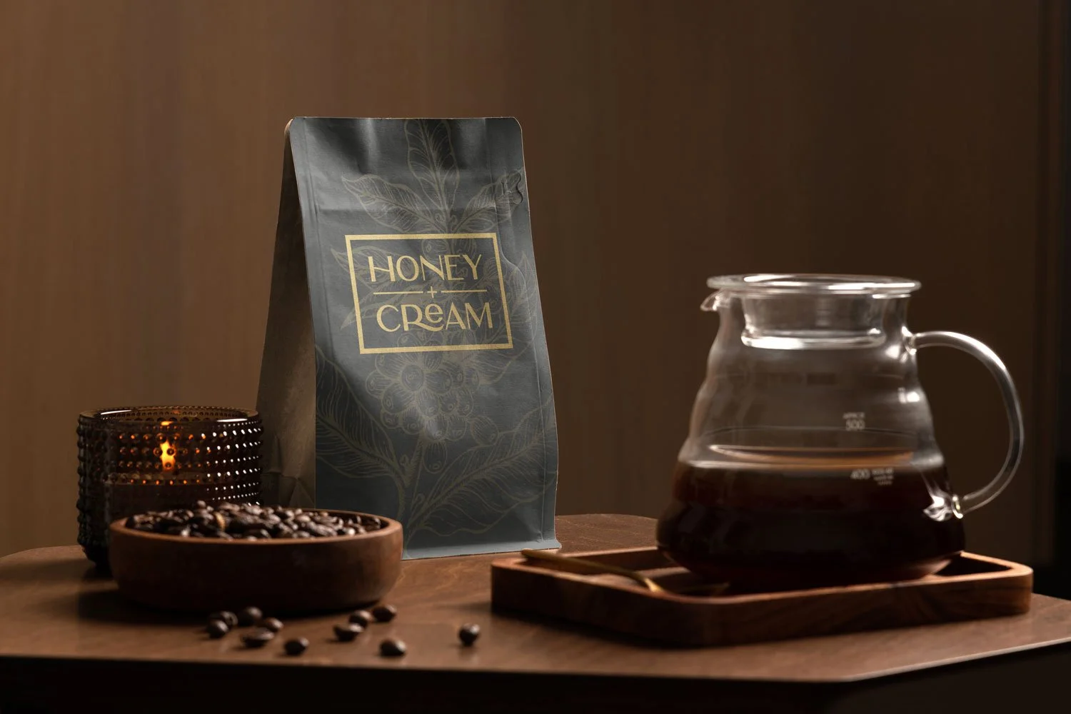 A bag of honey and cream coffee, a glass coffee pot with brewed coffee, coffee beans in a wooden bowl, and a candle holder on a wooden table.