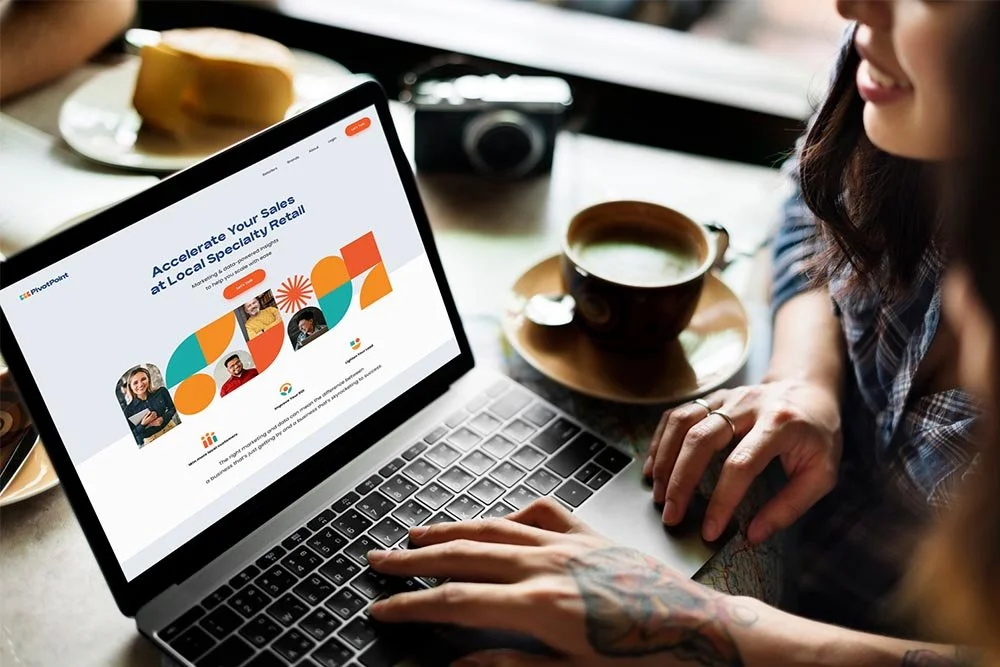 Two women look at a laptop screen displaying a webpage with the title 'Accelerate Your Sales at Local Specialty Recall' and colorful graphics. One woman has tattoos on her arm, and they are sitting at a table with coffee cups and plates with food.